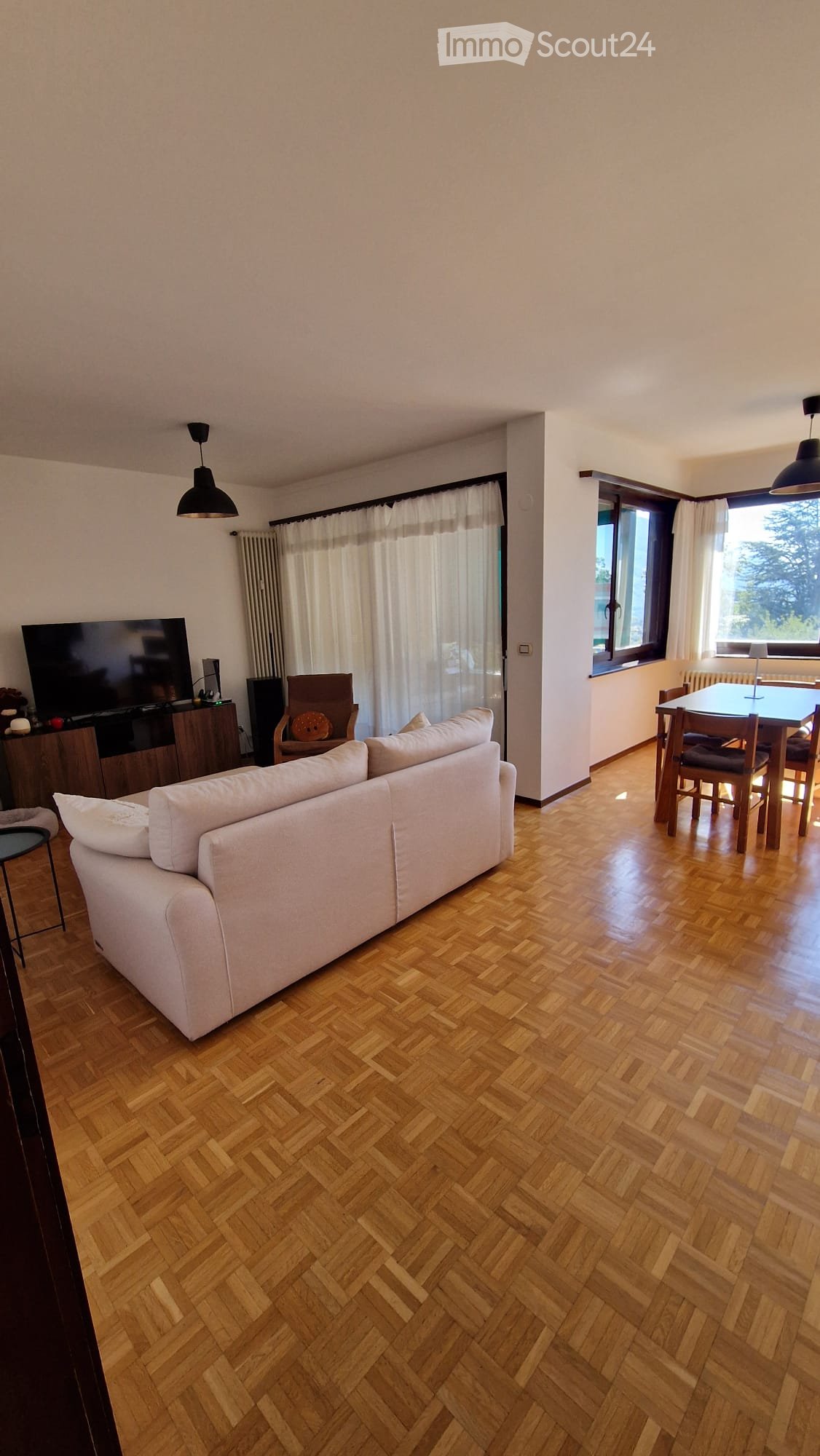 A spacious living and dining area with wooden herringbone flooring, large windows with white curtains, a white sofa, a wooden dining table with chairs, and a television on a wooden stand.