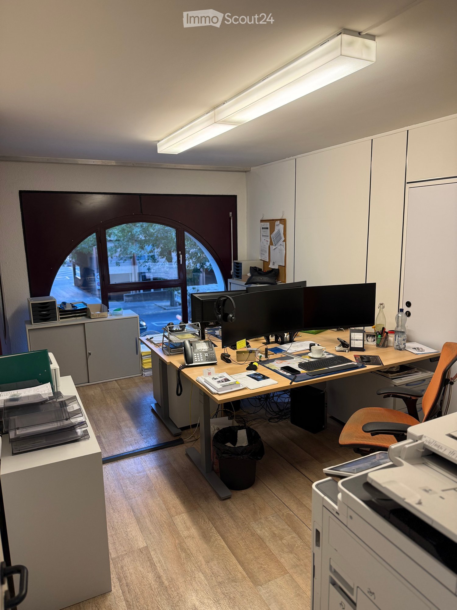 Office space with a wooden desk, ergonomic chair, dual monitors, keyboard, mouse, printer, telephone, and various office supplies.