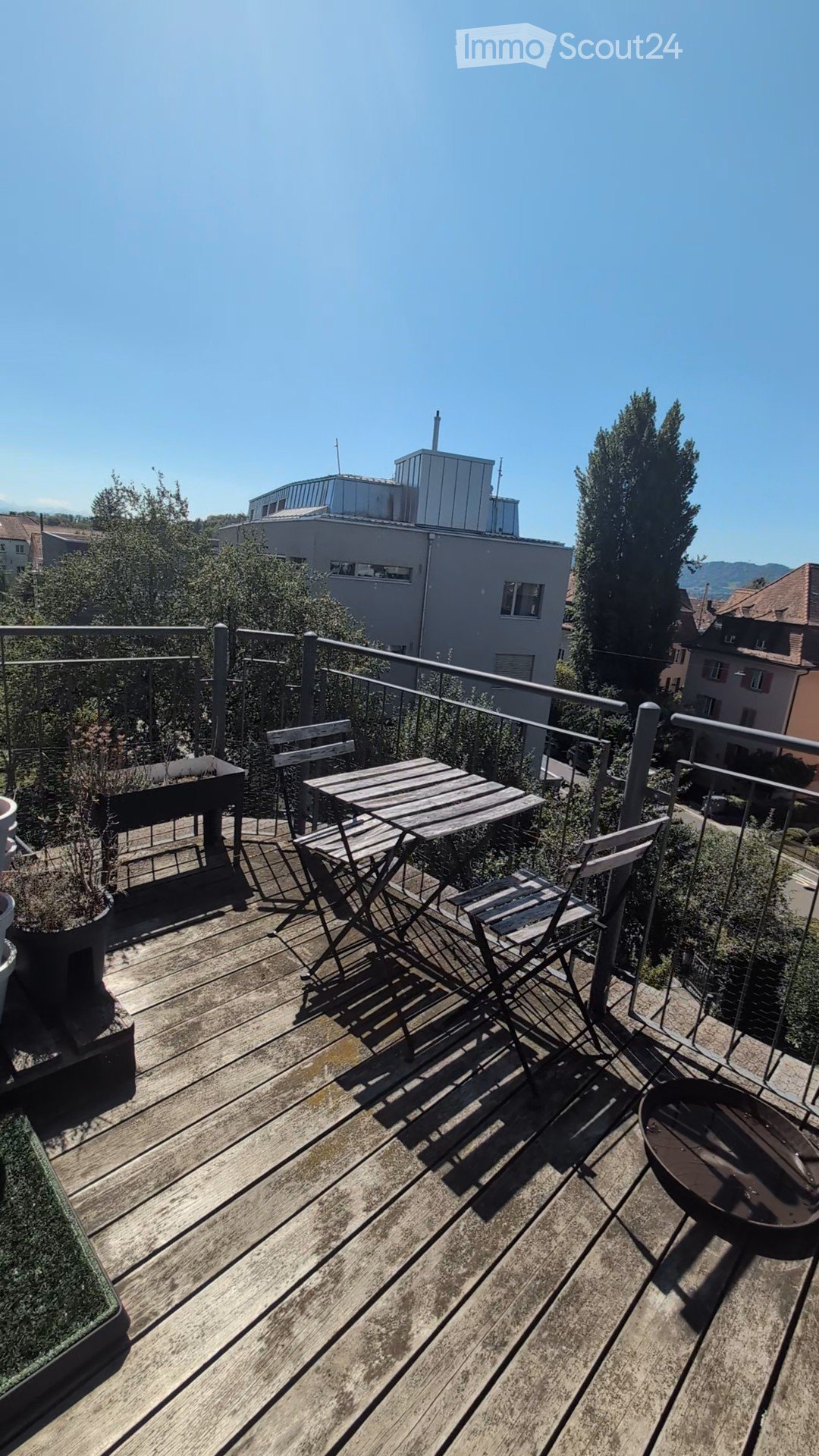 Wooden deck with a small table, chairs, potted plants, and a fire pit, overlooking a cityscape with buildings and trees.