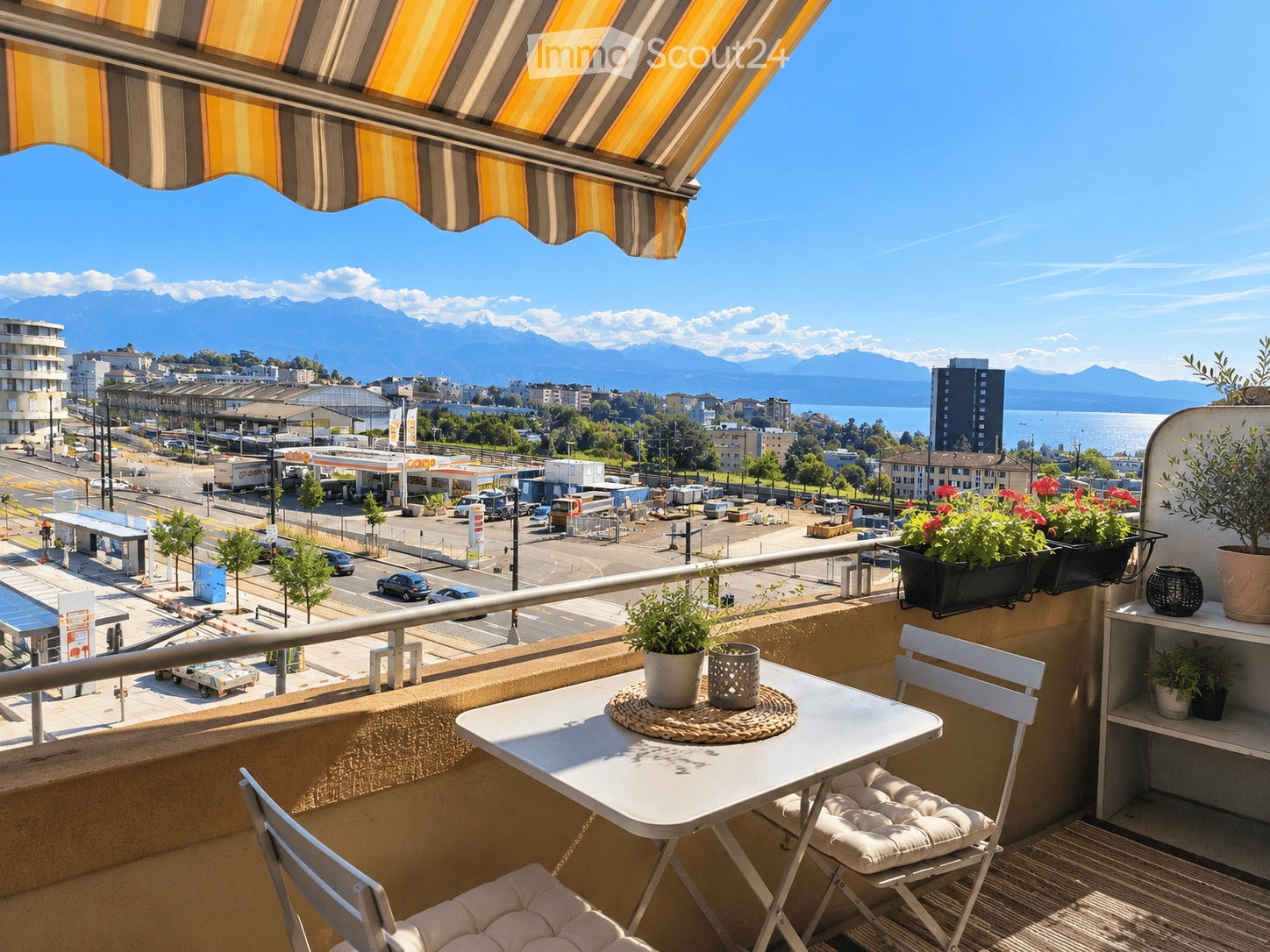Balcony with a table, chairs, and potted plants, overlooking a cityscape with mountains in the background.