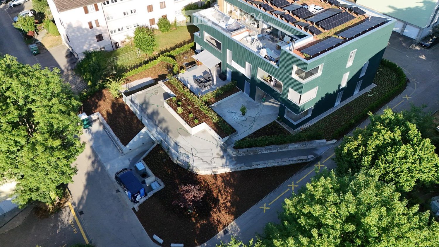 A modern green building with solar panels on the roof, surrounded by landscaped gardens and pathways. The building has multiple levels and large windows, with a raised ground floor and a parking area nearby.