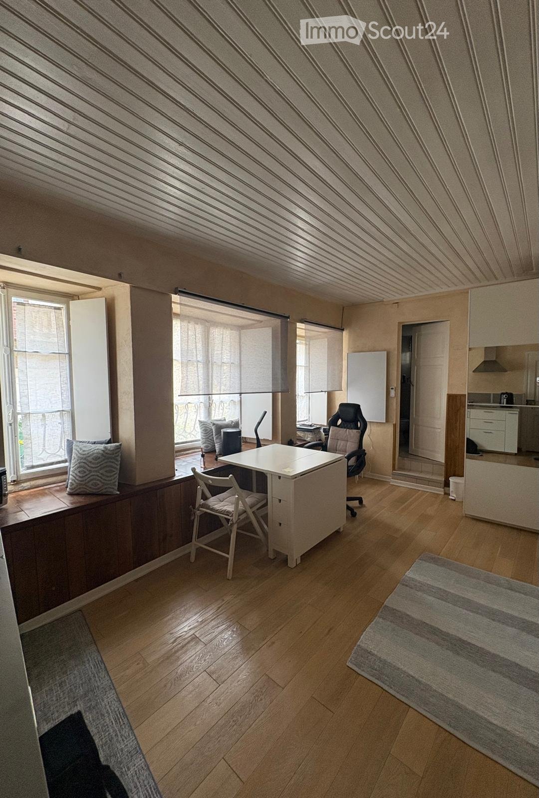 Bright, modern room with wooden flooring, large windows, a desk with chairs, a kitchen area, and a balcony view.