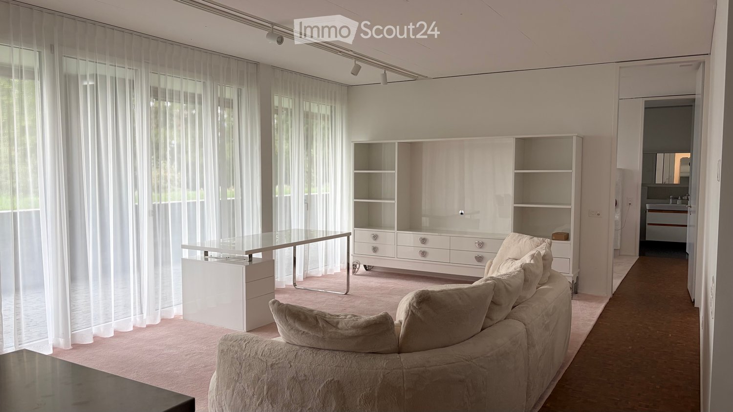Modern living room with a light pink carpet, white walls, large windows with sheer curtains, a beige sofa, a glass coffee table, and a white shelving unit.