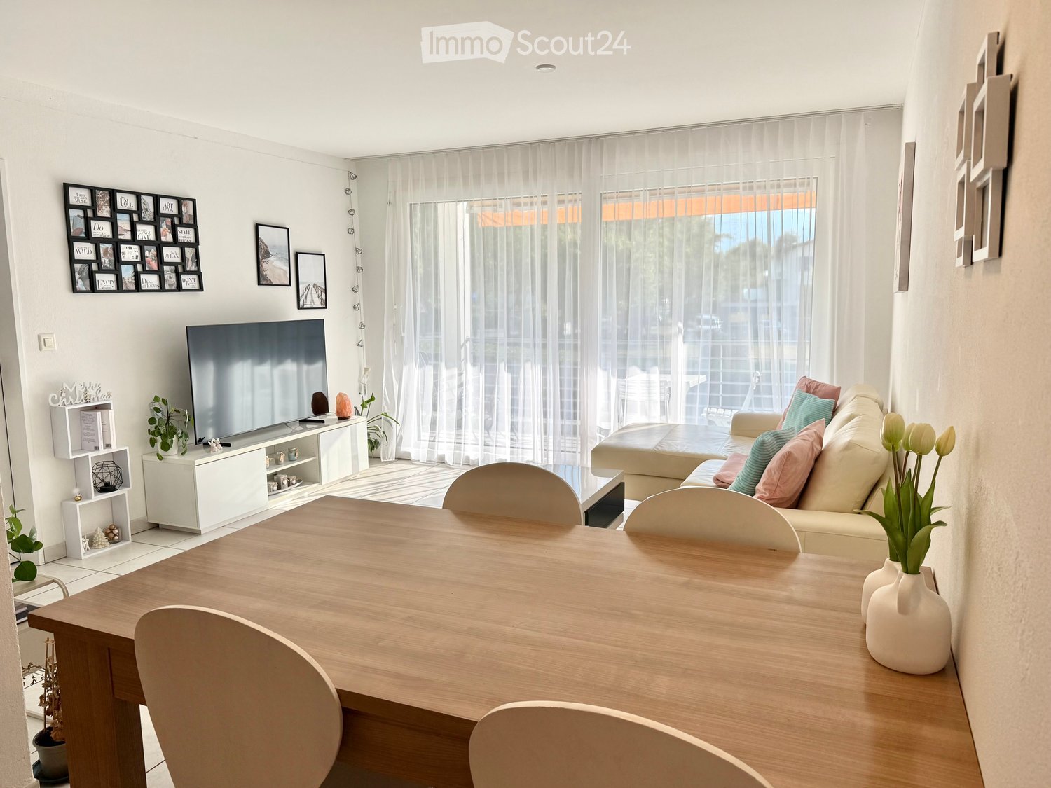 Bright living room with a beige sofa, wooden dining table, chairs, TV, and decorative shelves.