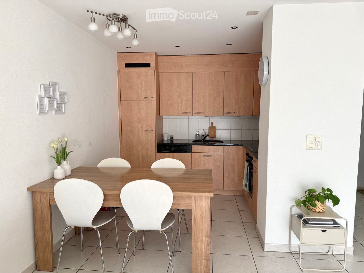 Modern kitchen with light wooden cabinets, a wooden dining table with four white chairs, a dishwasher, sink, and a small side table with a plant.