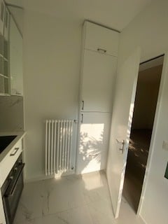 Empty room with white walls, tiled floor, radiator, and open door leading to another space.