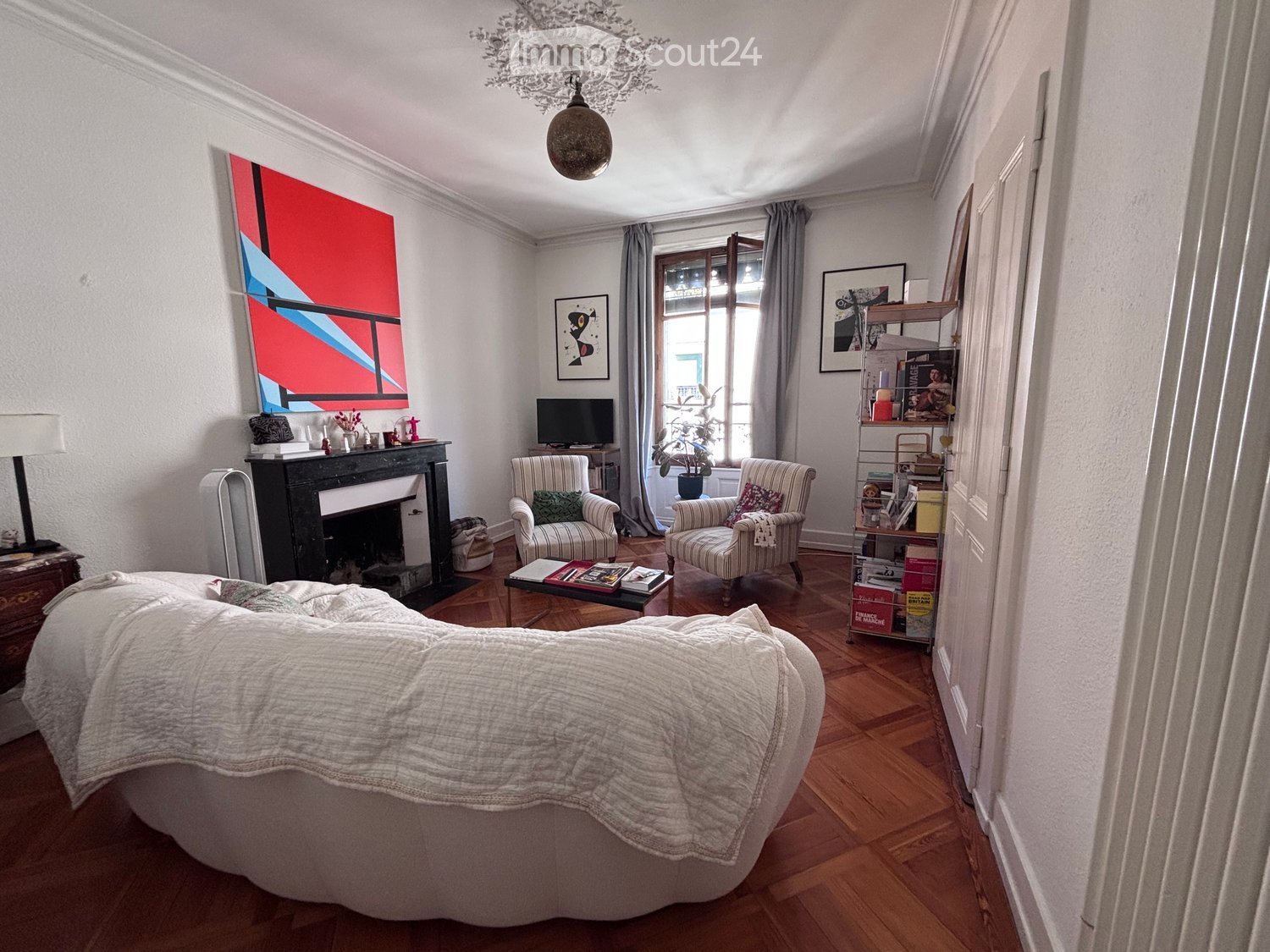 A cozy living room with a large sofa, two armchairs, a coffee table with books, a fireplace, a television, a shelf with books and decorative items, and a chandelier.