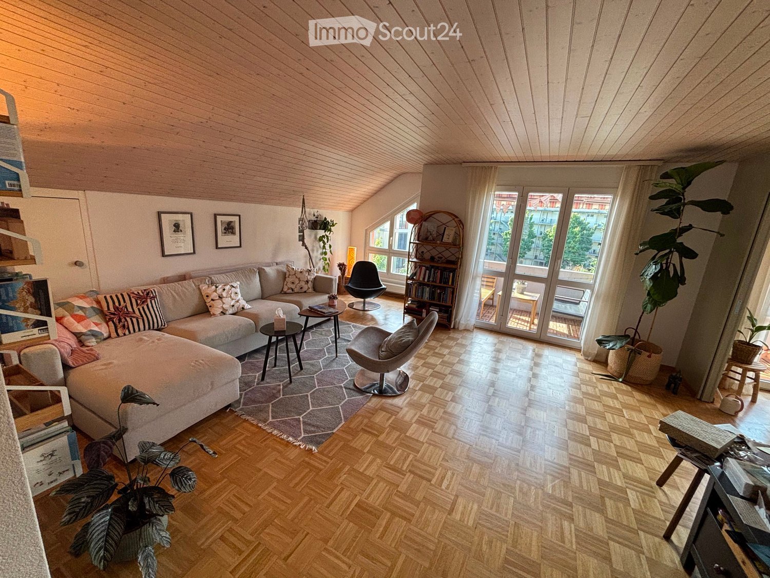 Modern living room with wooden floors, slanted wooden ceiling, large windows, and a balcony. Features a grey sectional sofa, coffee tables, chairs, bookshelf, and plants.
