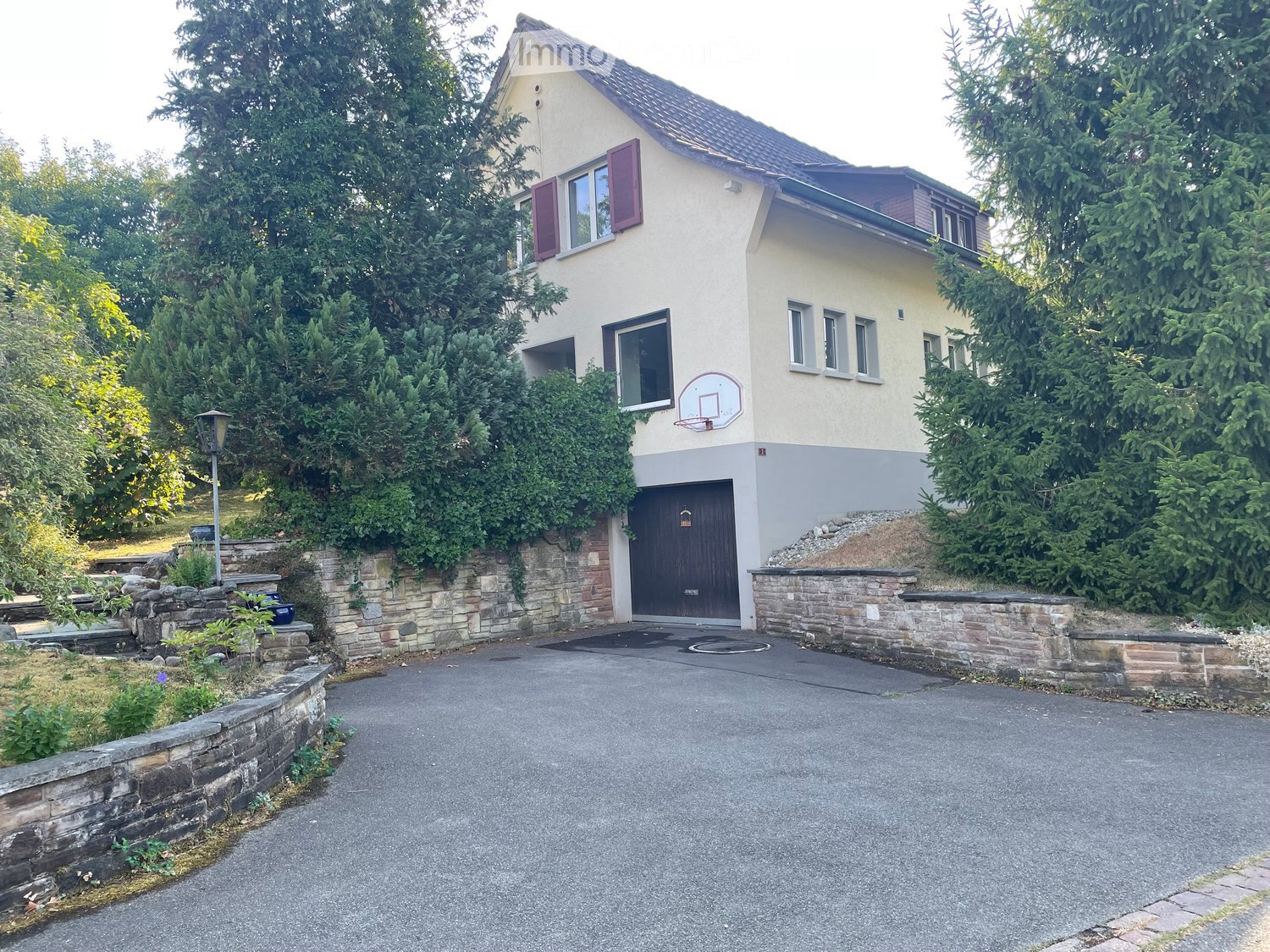 Two-story house with beige exterior, purple shutters, garage, basketball hoop, and stone retaining walls.
