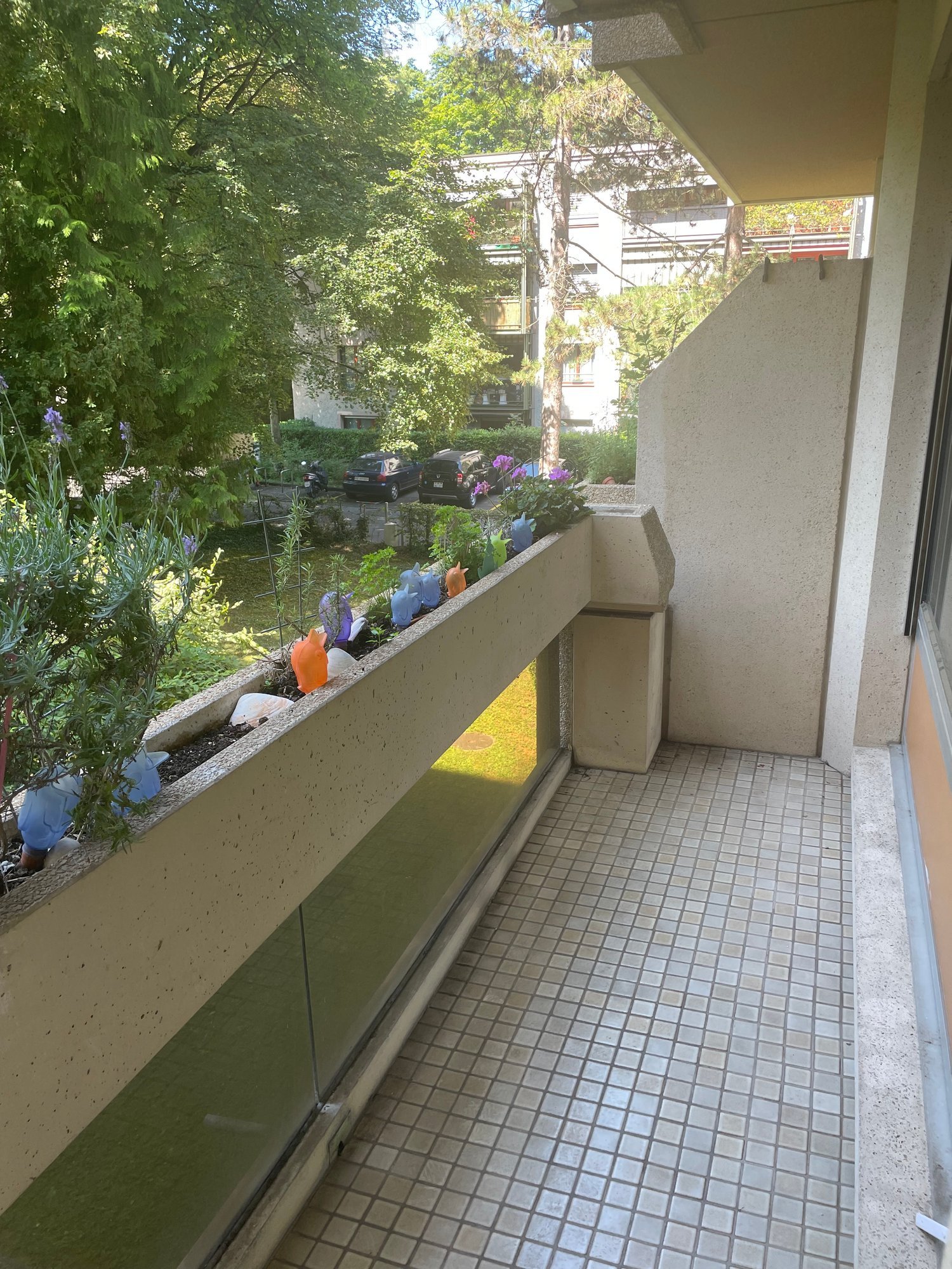 Balcony with tiled floor, small garden area with plants and decorative items, overlooking a green space with trees and parked cars.