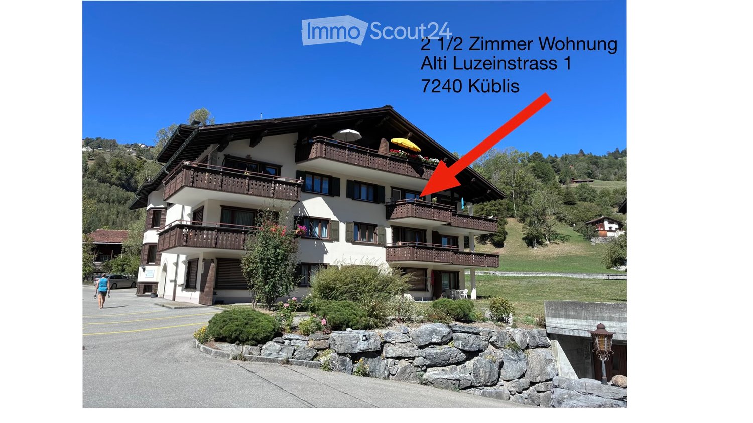 A multi-story residential building with balconies, located in a scenic, hilly area with greenery and a clear blue sky.