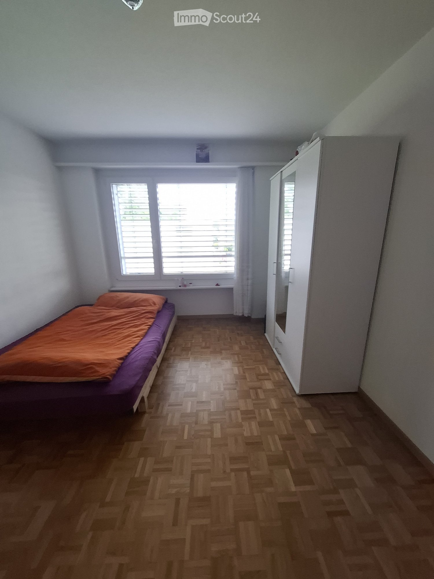 Bedroom with wooden herringbone flooring, a single bed with orange and purple bedding, a white wardrobe, and a window with blinds.
