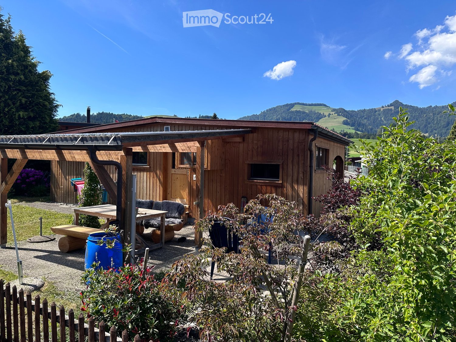A wooden cabin surrounded by lush greenery and mountains, with a small outdoor seating area featuring a table, sofa, and bench.