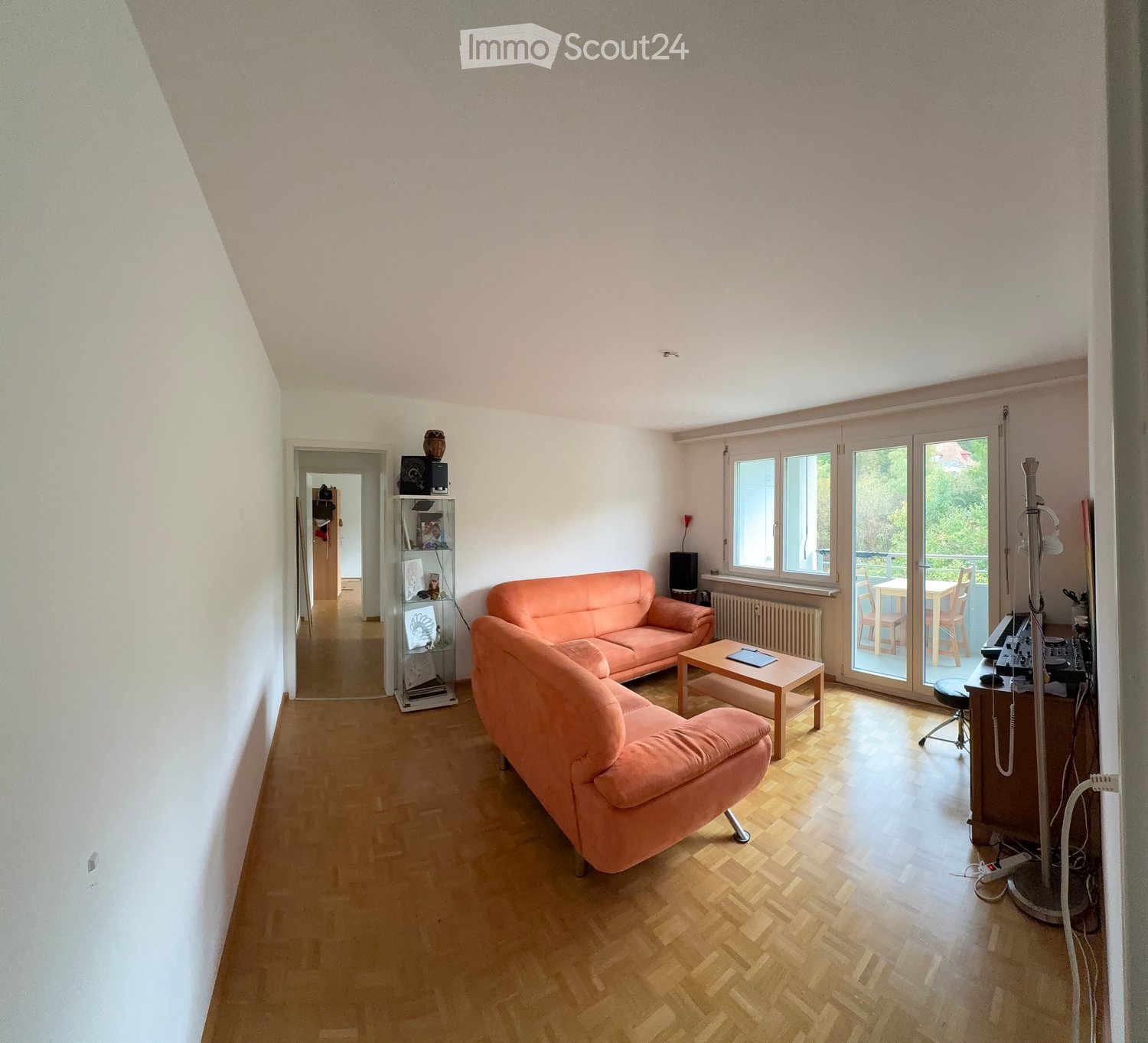 Bright living room with wooden flooring, large windows, and a balcony. Features a pink sofa, coffee table, and a small workspace area with a chair and table.