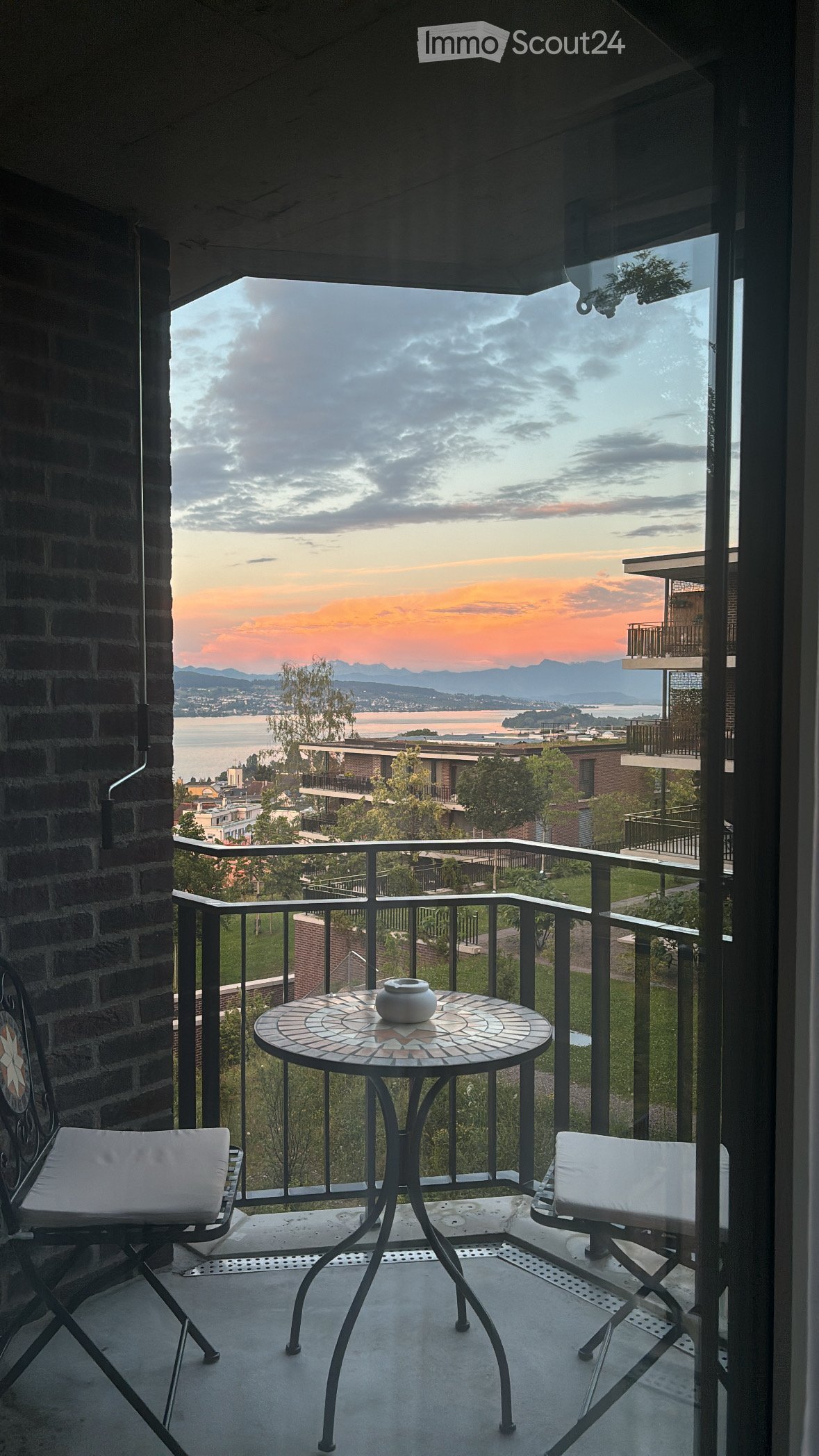 Balcony with a round mosaic table, two white cushioned chairs, and a decorative vase, overlooking a scenic view of a lake and distant mountains.