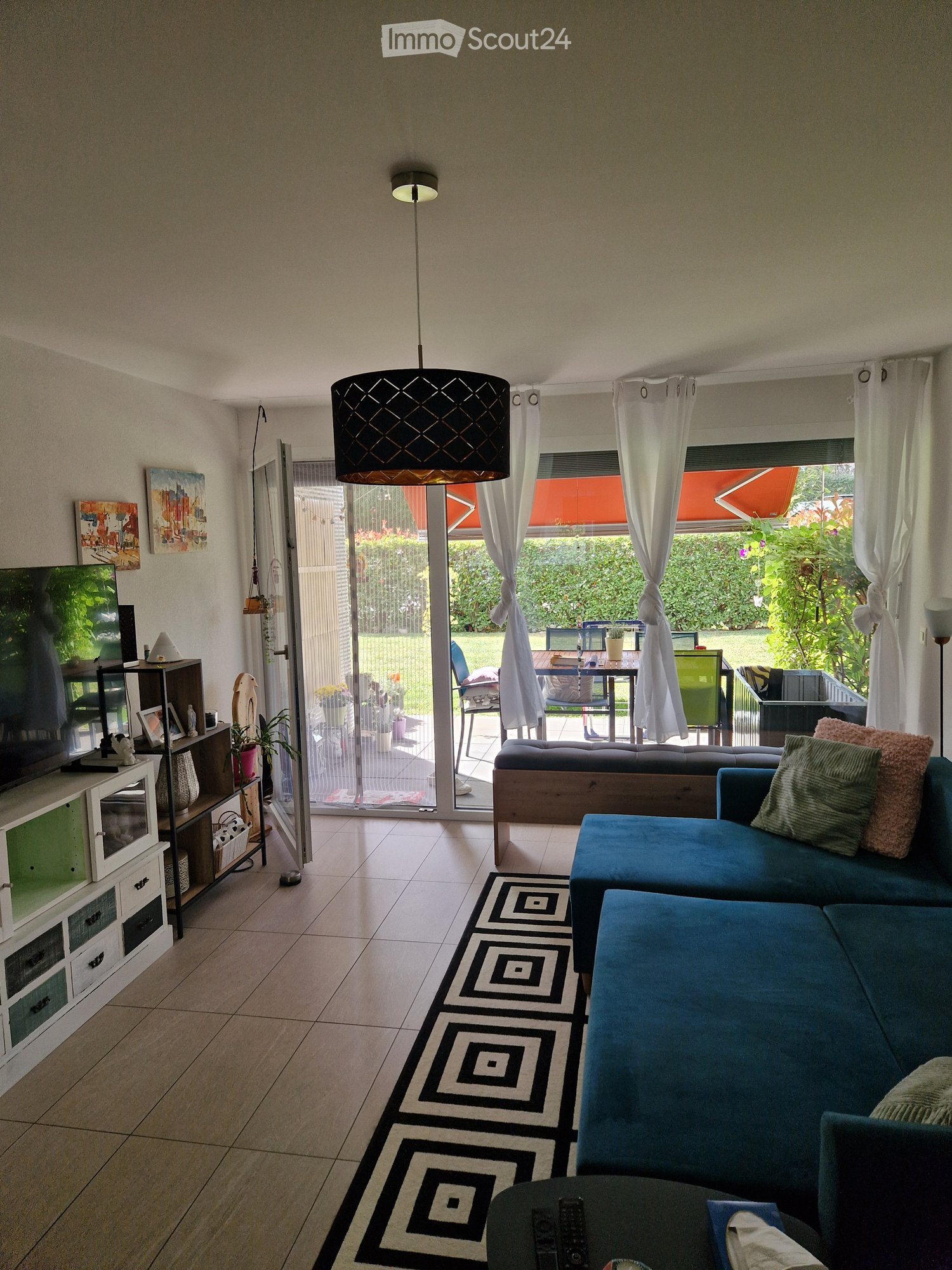 Modern living room with a blue sectional sofa, coffee table, TV on a shelf, pendant light, and an open sliding door leading to a patio with outdoor furniture.
