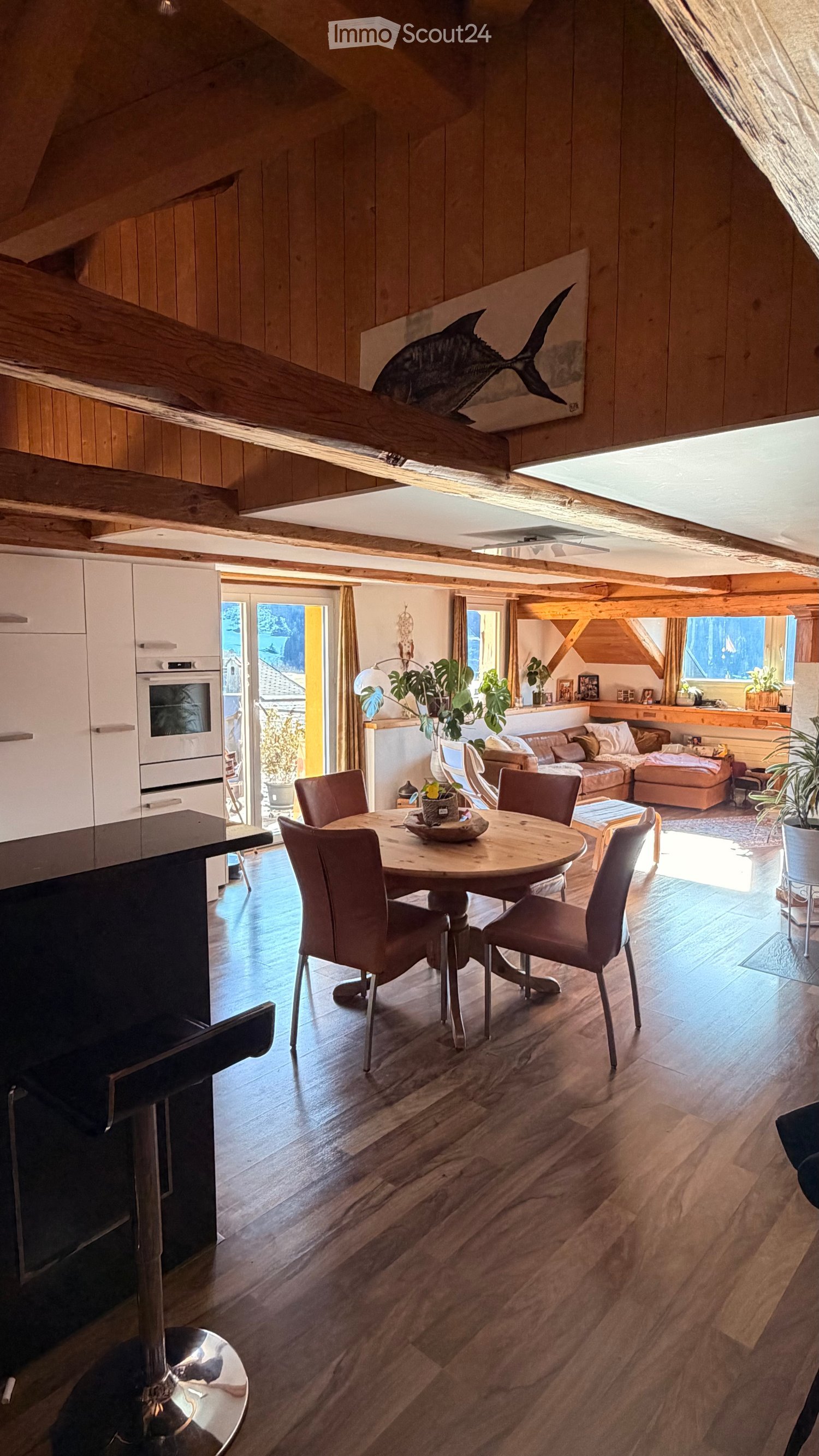 Open-plan living and dining area with wooden floors, exposed wooden beams, a round dining table with six chairs, a sofa with an ottoman, kitchen cabinets, and a modern oven. Large windows and a sliding glass door lead to a balcony.