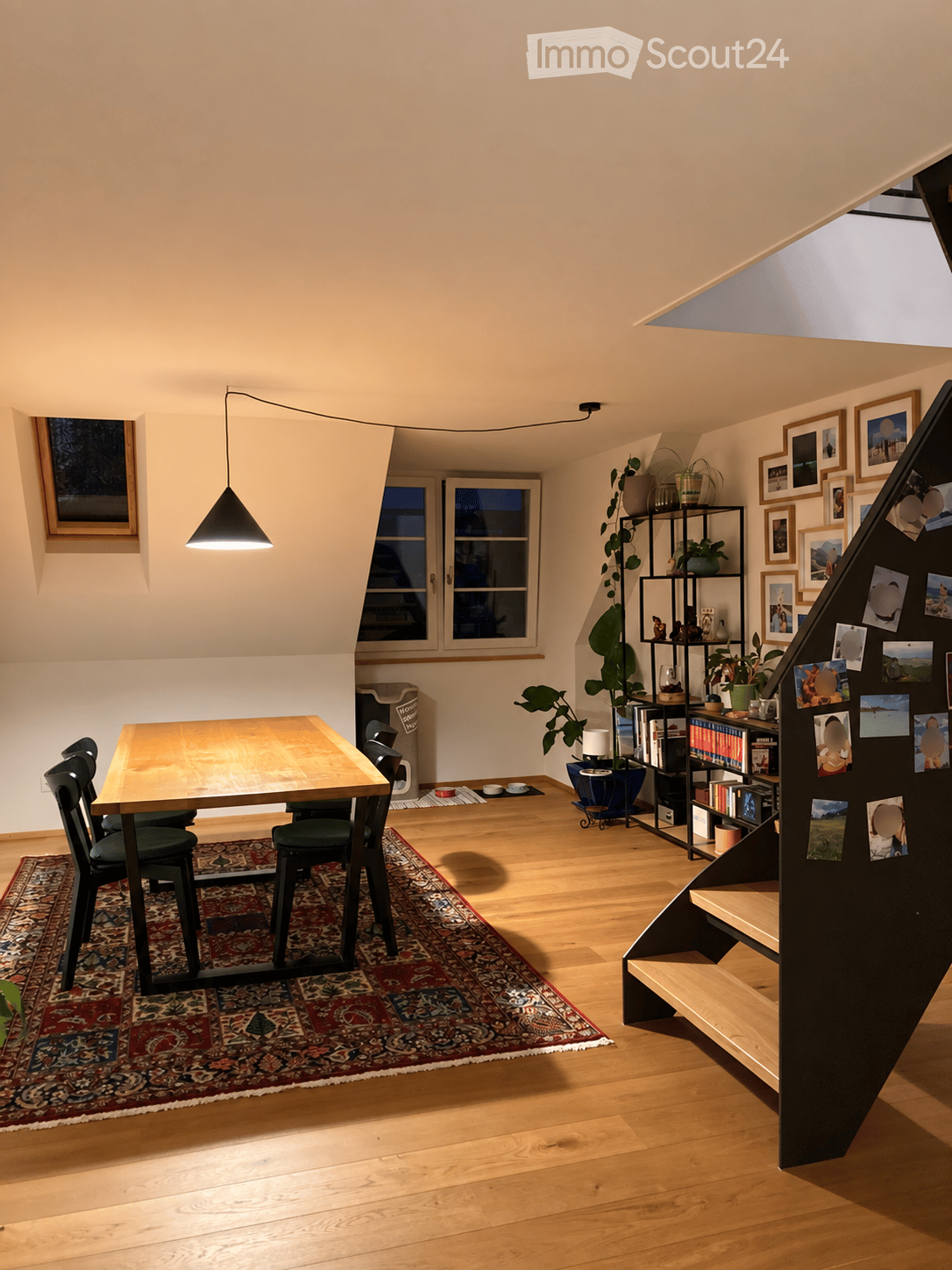 Bright, open room with wooden flooring, a dining table with chairs, a large plant, a bookshelf, and a staircase leading upwards.