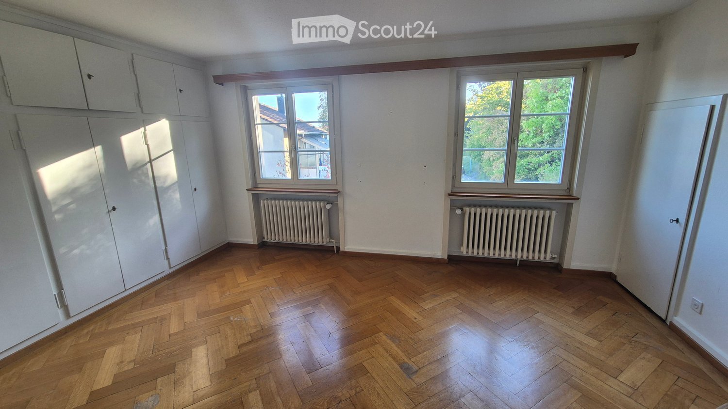 Empty room with wooden herringbone flooring, white walls, built-in wardrobes, and two windows with radiators beneath them.