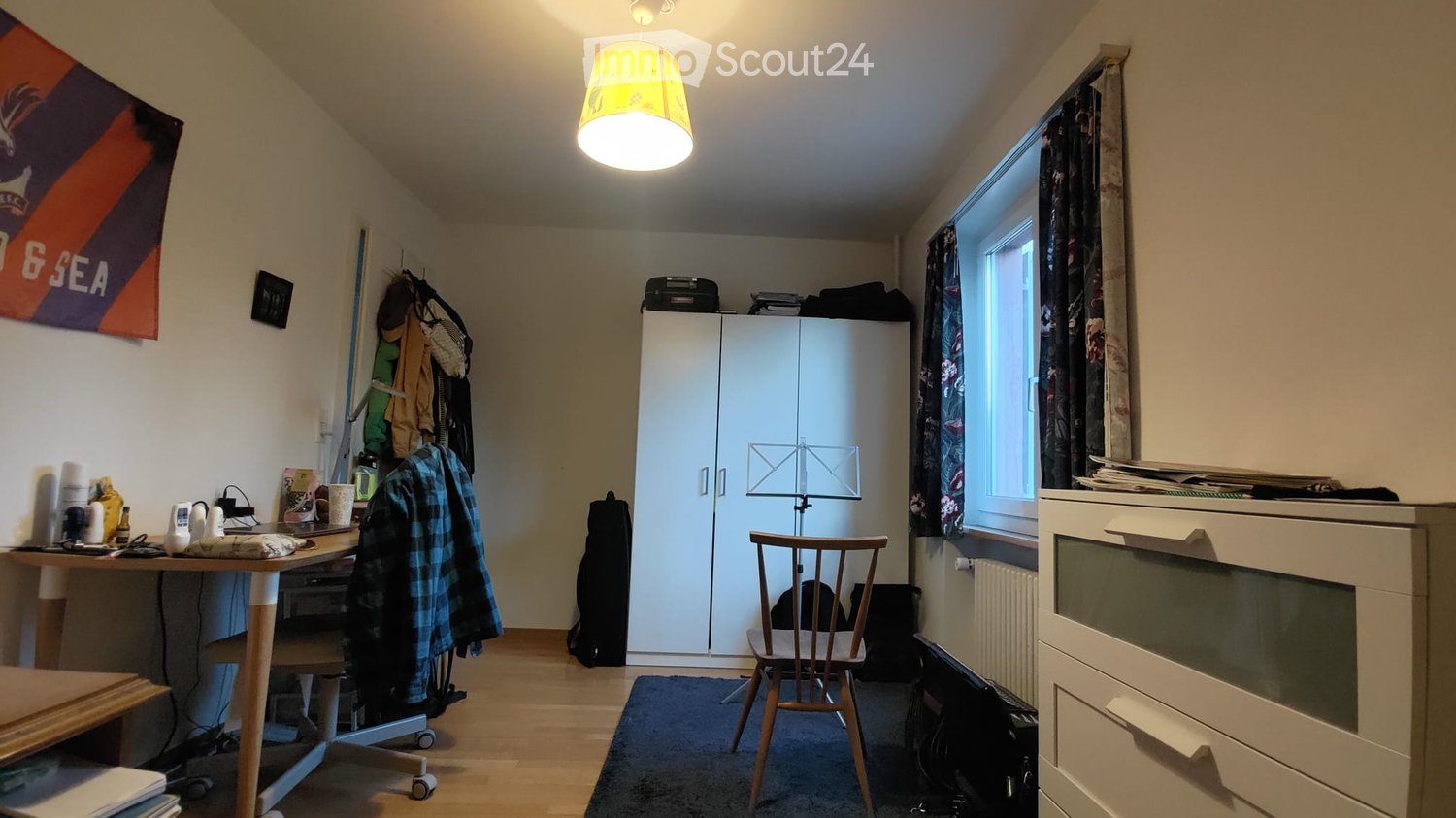A furnished room with a wooden floor, a large window with curtains, a white cabinet, a desk with a laptop, a chair, and a lamp hanging from the ceiling.