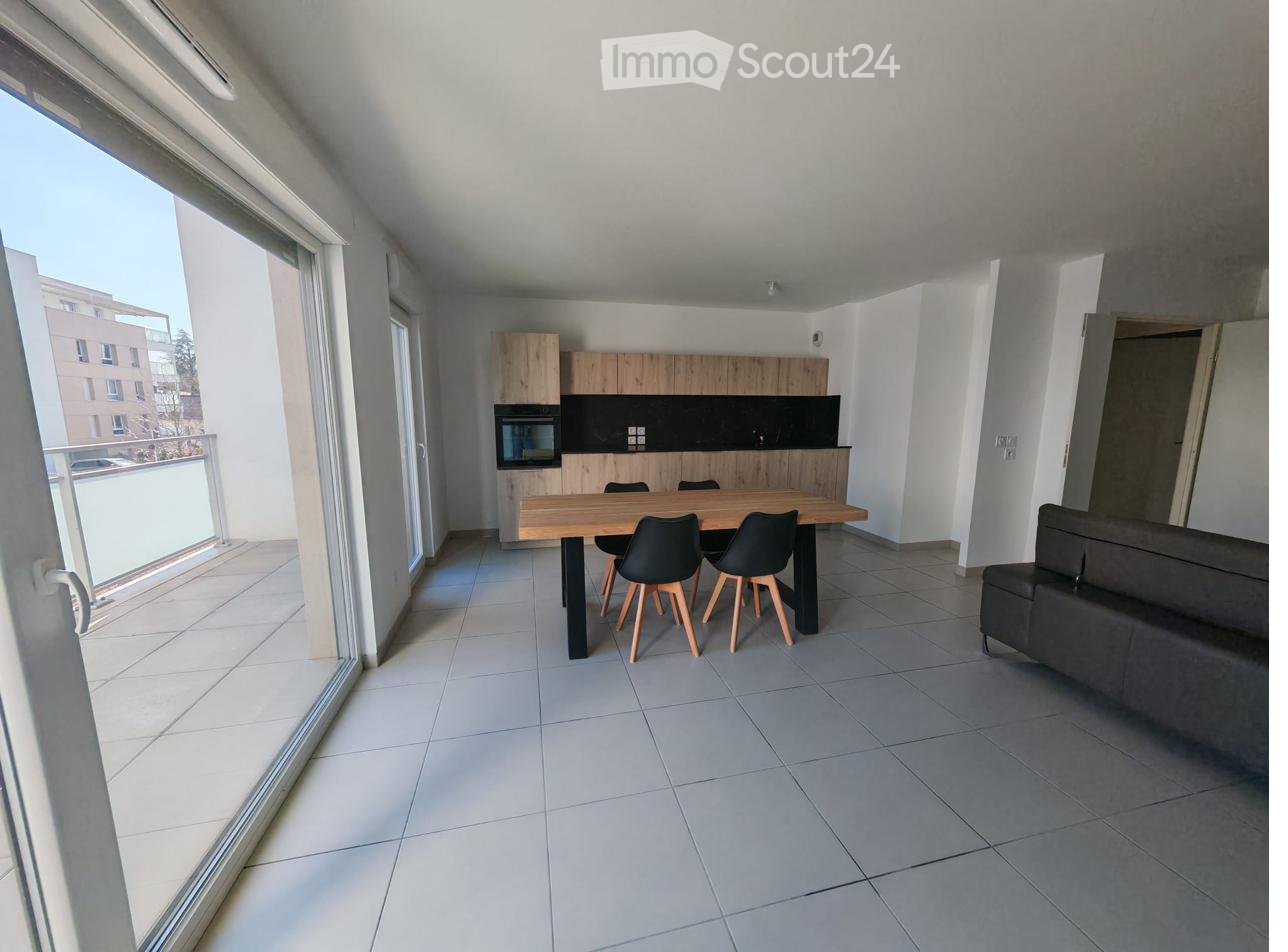 Modern open-plan living and dining area with a grey sofa, wooden dining table, four chairs, and a kitchen area with cabinets and an oven. Large windows and a balcony are visible.