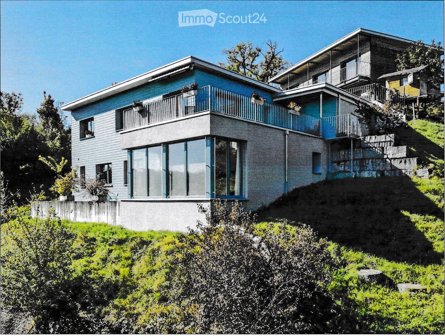 Modern two-story house with blue and grey exterior, large windows, balconies, and built on a hillside with surrounding greenery.