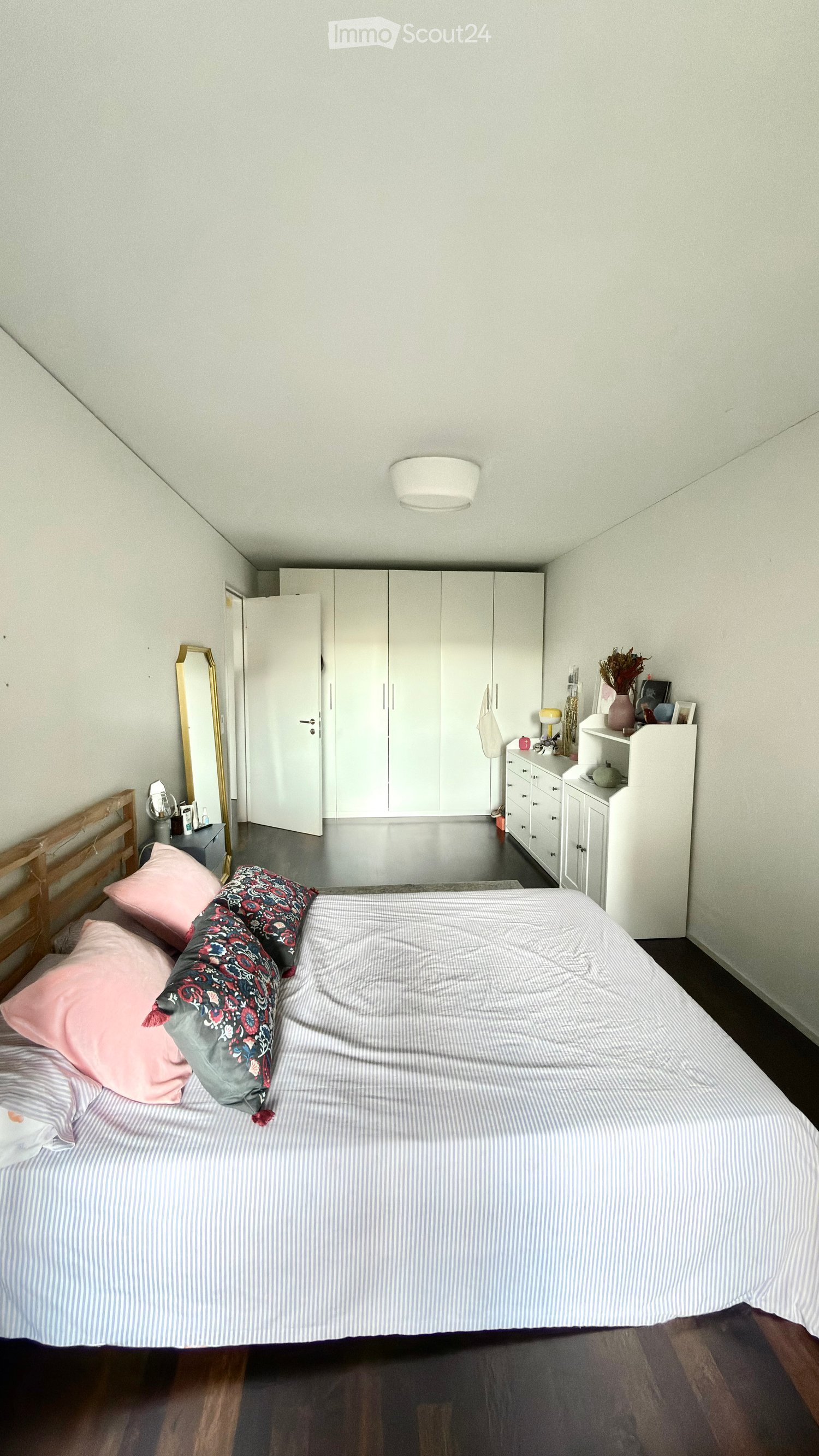 A modern bedroom with a large bed, striped bedding, pink and patterned pillows, a wooden headboard, white wardrobe, a gold-framed mirror, and a nightstand with a lamp and decorative items.
