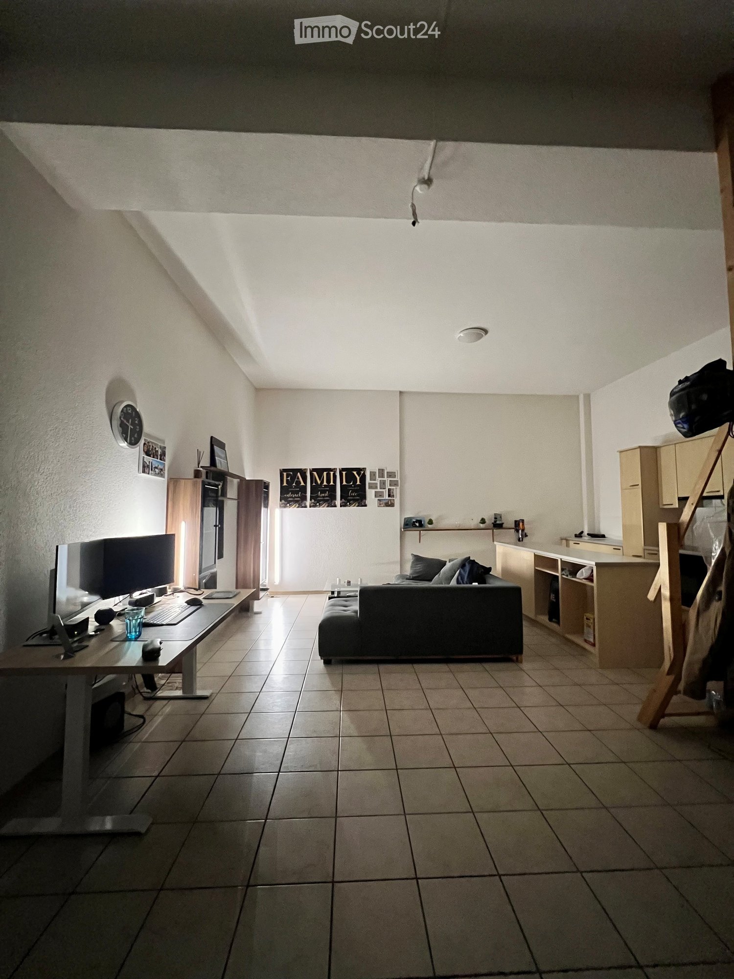 A modern living room with a sofa, a table with a screen and keyboard, a chair, and a cabinet. The room has tiled flooring and white walls.