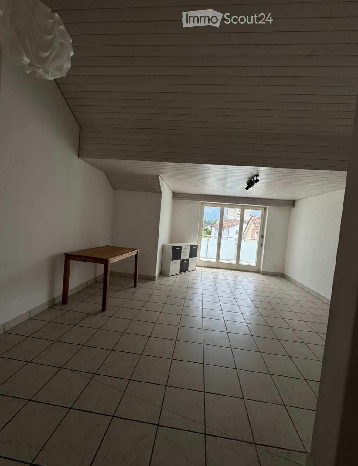 A small, unfurnished room with tiled flooring, a wooden table, and a small cabinet near a large window with a view of the outside.