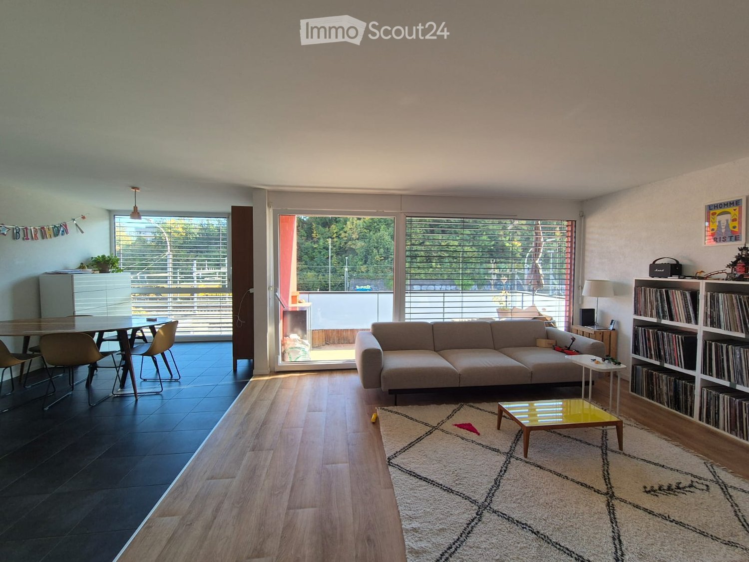 Modern living room with a sofa, coffee table, dining table, chairs, bookshelf, and a lamp. Large windows and a balcony are visible.