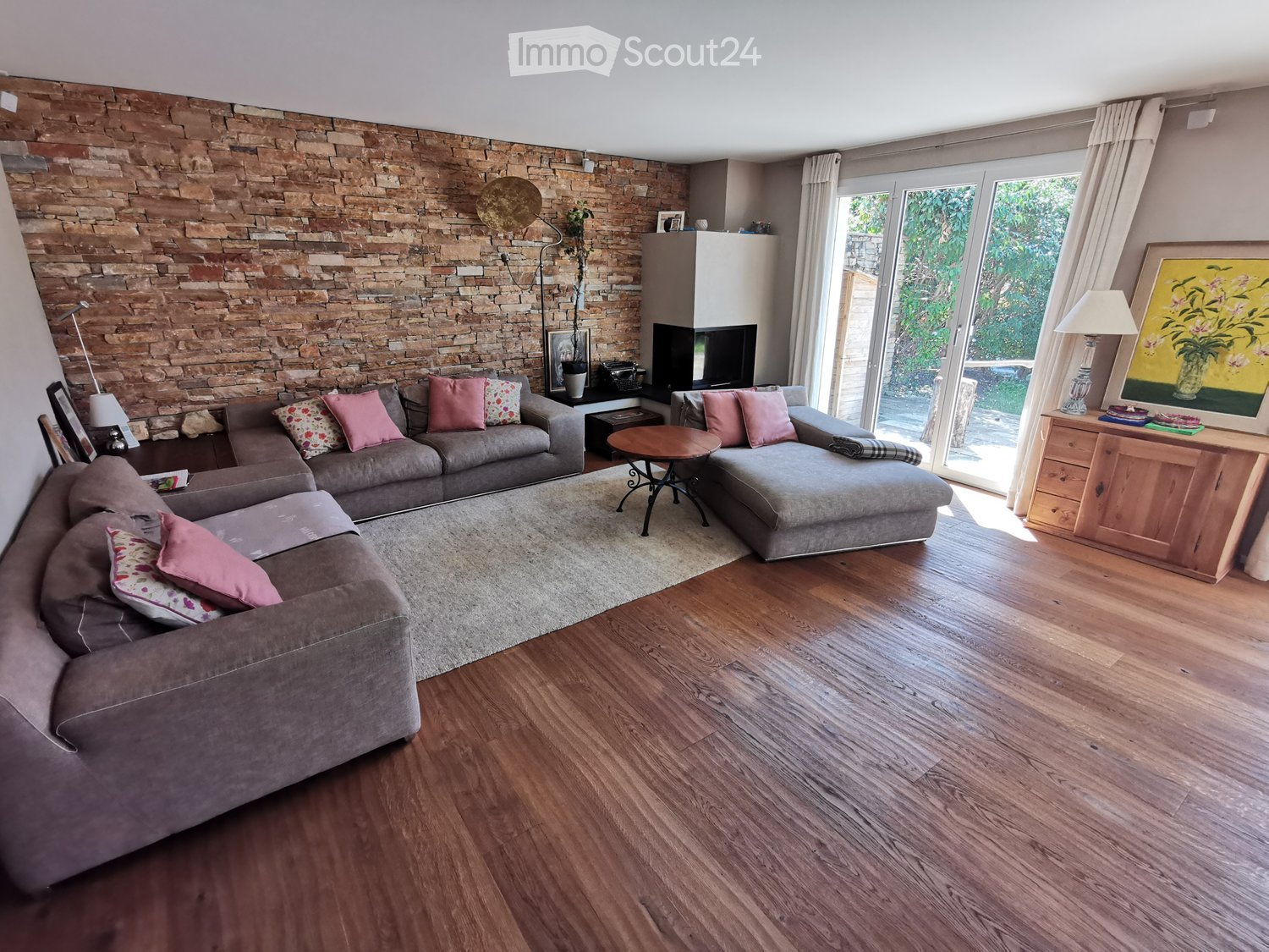 Modern living room with a stone accent wall, grey sectional sofa, coffee table, side tables, floor lamp, fireplace, and large windows with curtains.