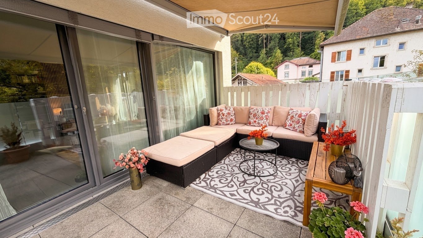 Balcony with a modern seating area, including a sectional sofa, small round table, and decorative vases with flowers. The space features a patterned rug and is enclosed by a wooden fence.