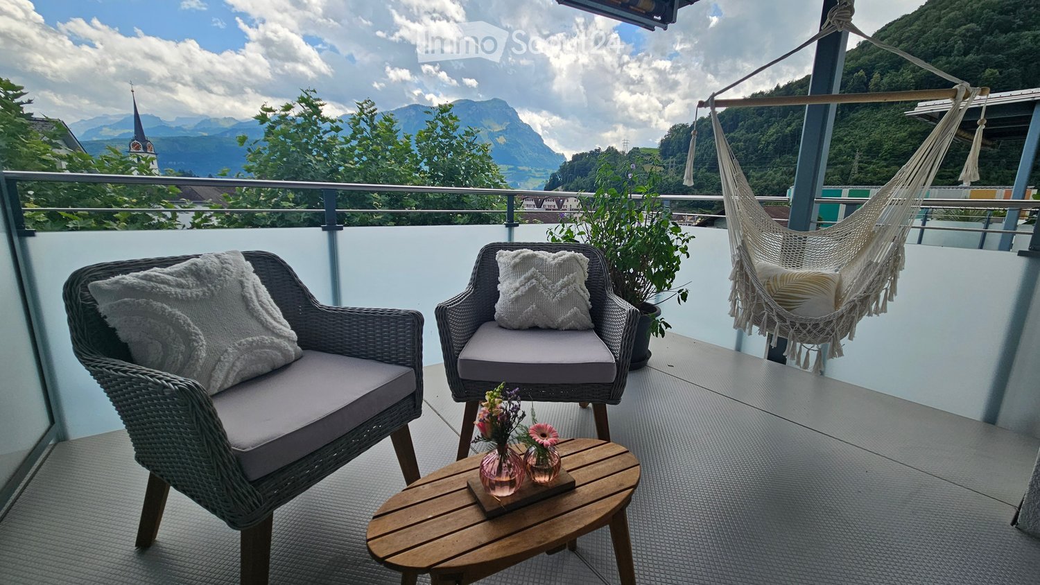 Balcony with two wicker chairs, a wooden coffee table, a hammock, and a potted plant, overlooking a scenic mountain view.