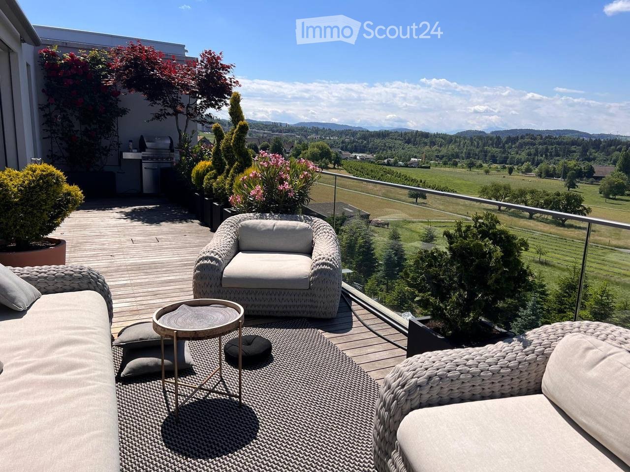 A spacious balcony with a view of a green landscape, featuring a wicker sofa, a wicker chair, a small round table, and several potted plants.