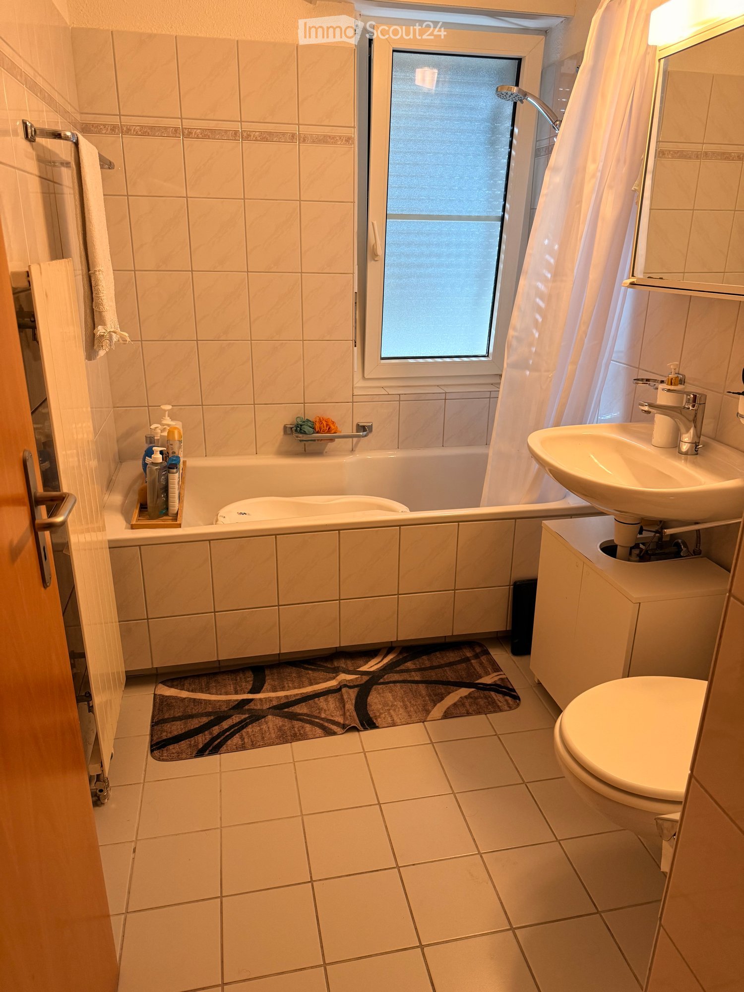 Bathroom with tiled walls and floor, bathtub, sink, toilet, mirror, and a towel on a rack. A small window above the bathtub provides natural light.
