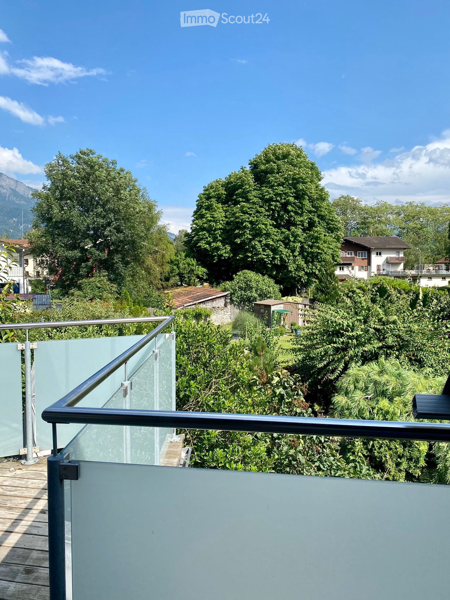 Balcony with glass railing overlooking a lush green garden and distant mountains under a clear blue sky.