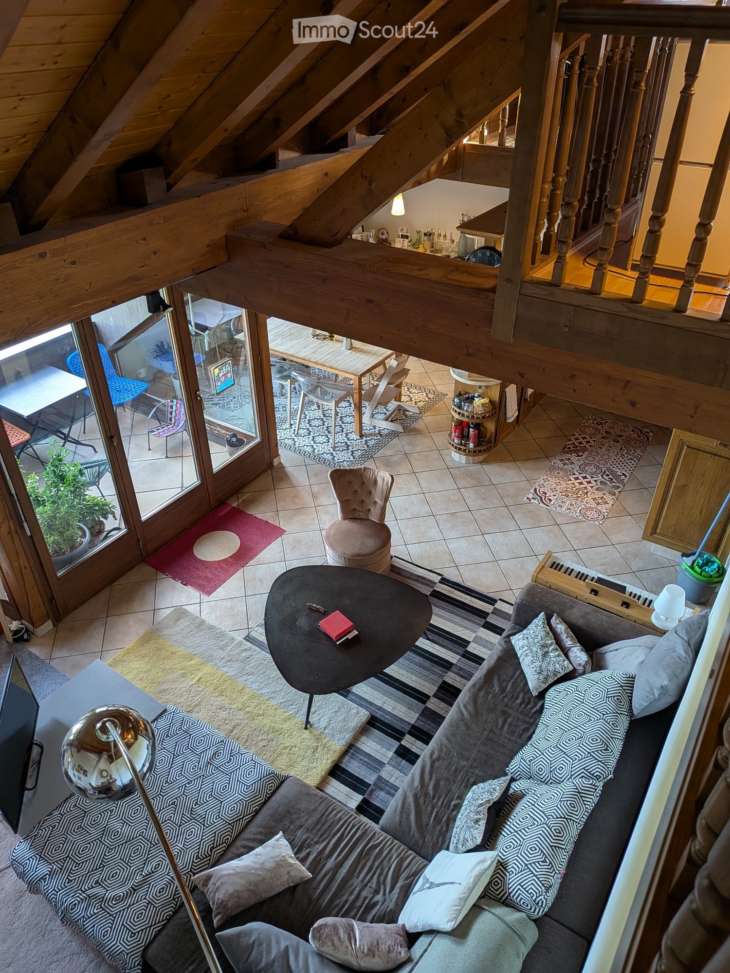 Living room with a grey sectional sofa, coffee table, small round stool, dining table, chairs, and a lamp. The room features wooden beams on the ceiling and a staircase leading to an upper level.