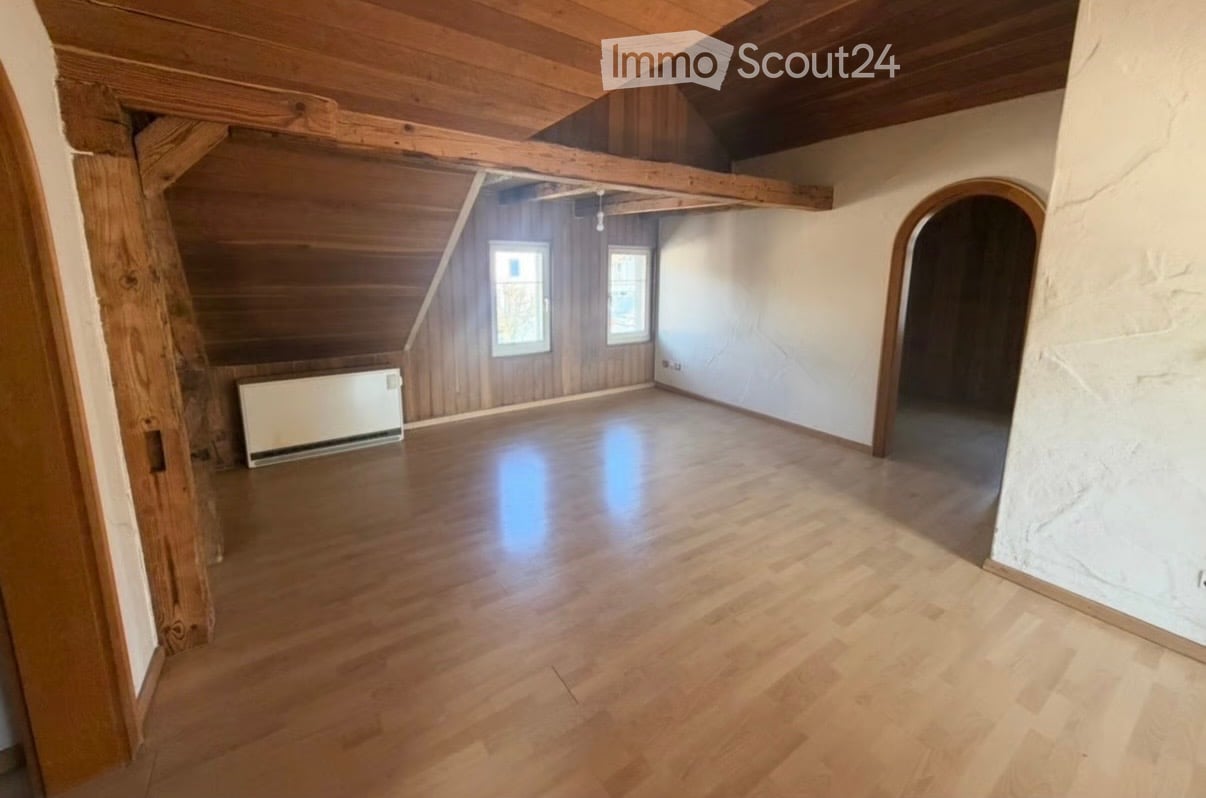 Empty attic room with wooden floors, exposed wooden beams, and two windows. The walls are painted white, and there is a radiator on the left side.