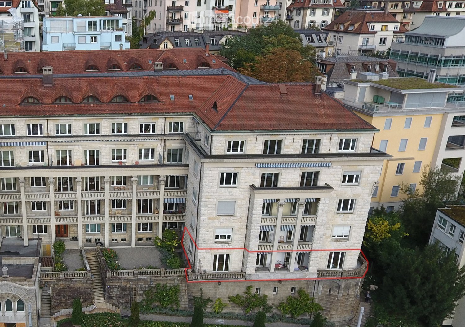 A multi-story residential building with a red-tiled roof, white stone facade, and multiple windows and balconies. The building is situated in a dense urban area with neighboring structures visible in the background.