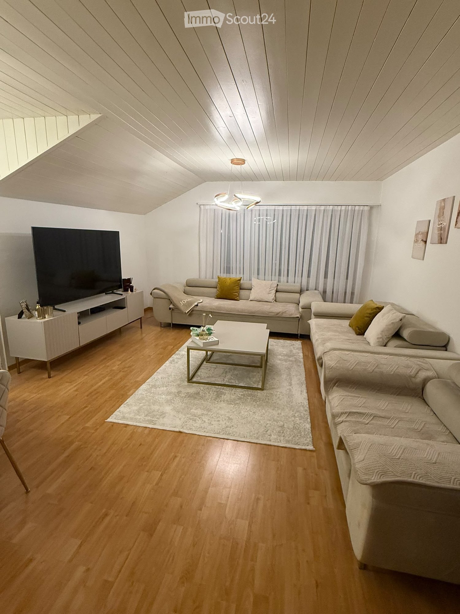 Modern living room with light wooden flooring, white walls, and a slanted ceiling. Features a large sectional sofa, coffee table, TV stand, and decorative pillows.