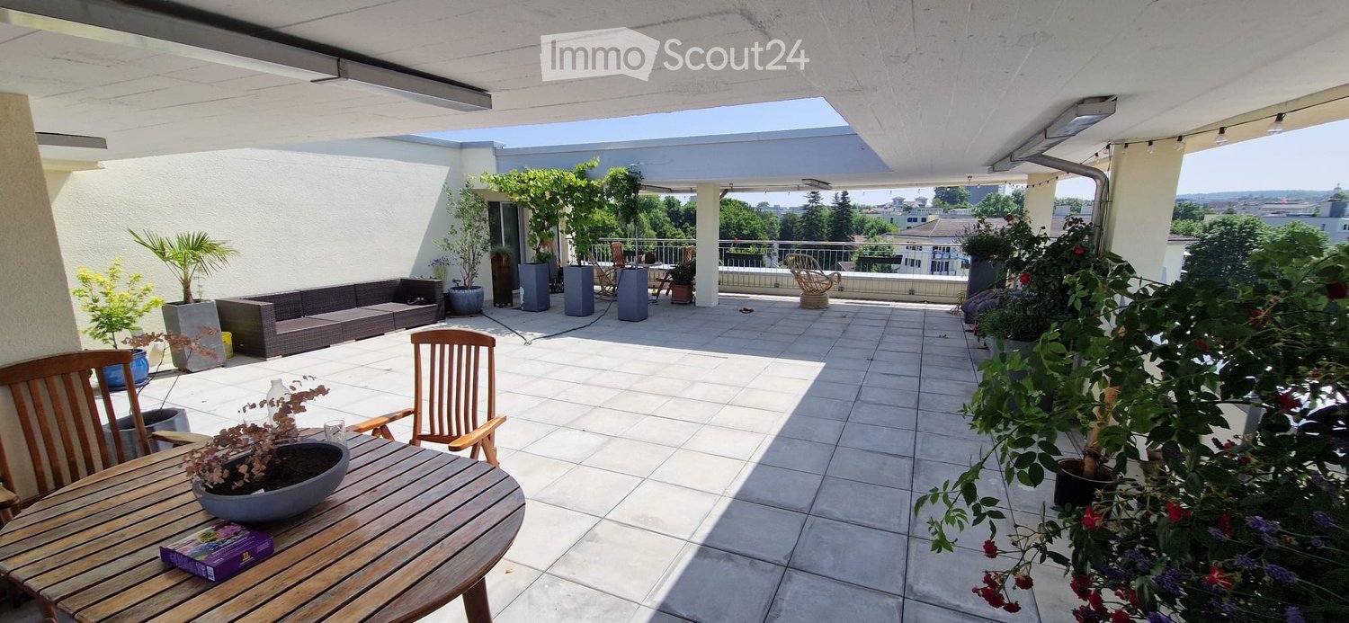 Spacious balcony with tiled floor, wooden table and chairs, sofa, potted plants, and scenic city view.