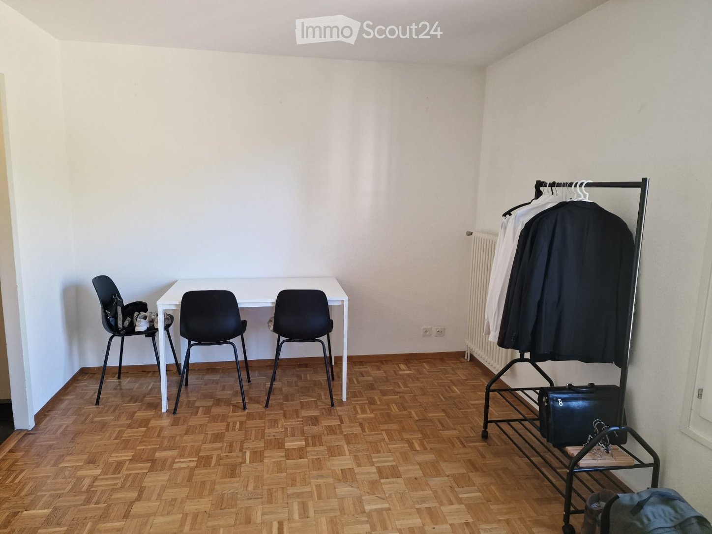 A small, unfurnished room with wooden herringbone flooring, white walls, a white table with three black chairs, a coat rack with jackets, and a radiator on the wall.