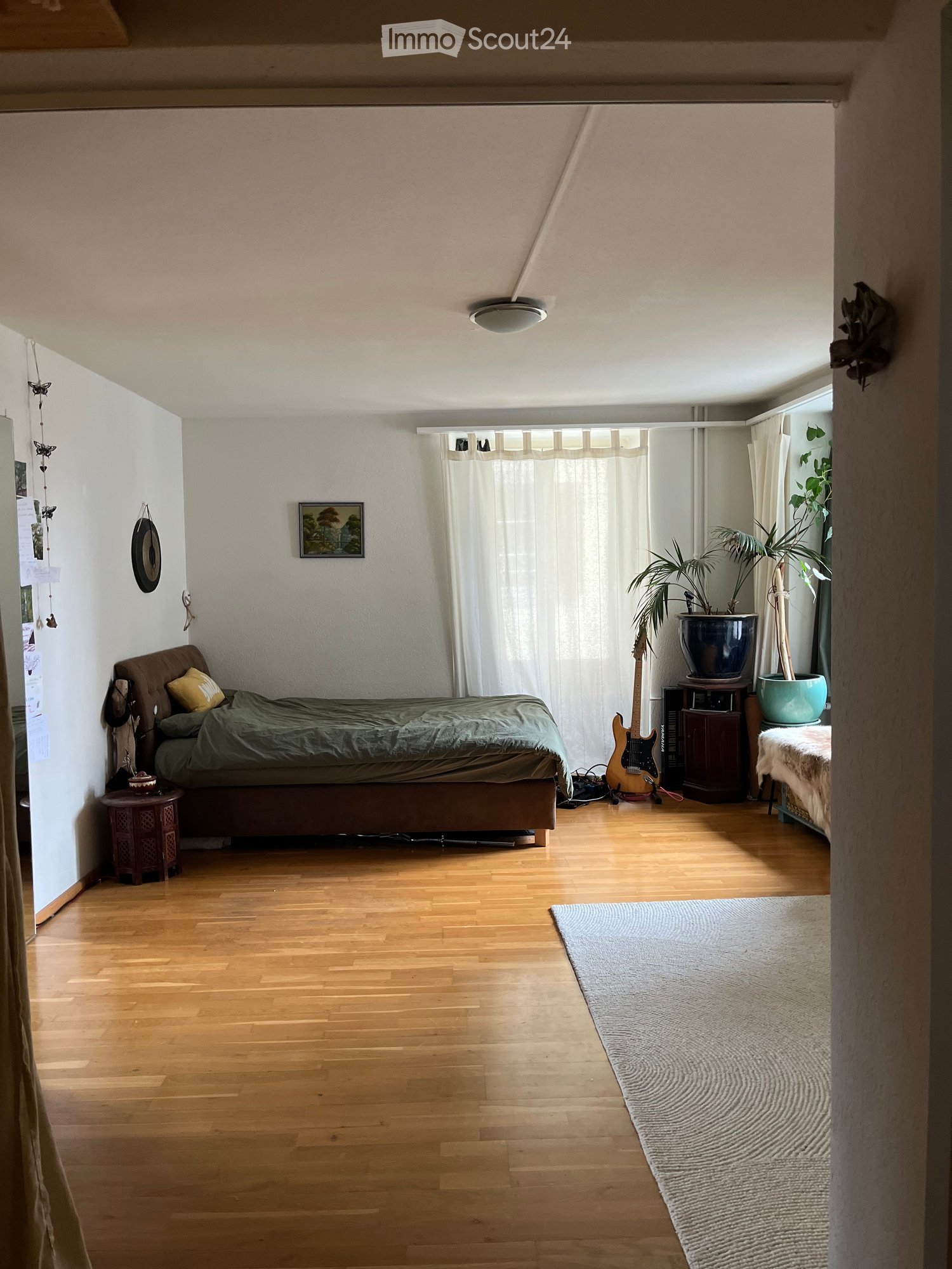 Cozy bedroom with wooden flooring, a bed with green bedding, a guitar leaning against the wall, a nightstand, a small table with a plant, and a large window with white curtains.