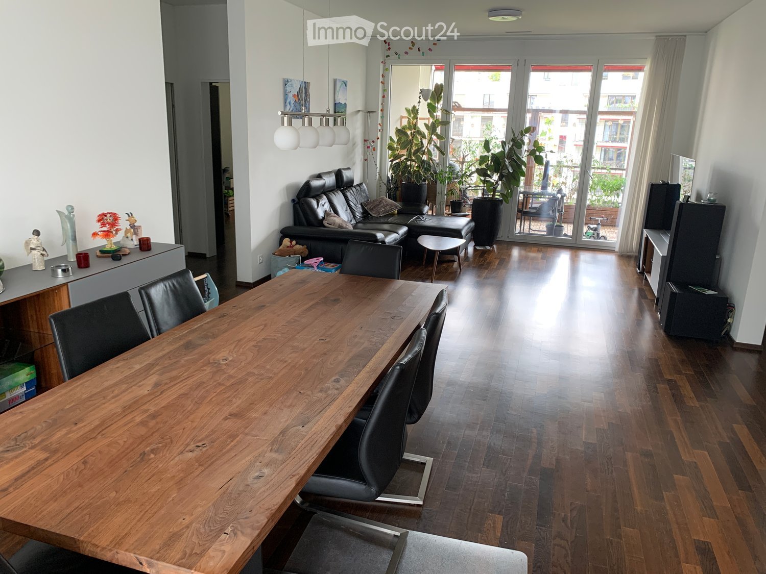 Bright, open living and dining area with wooden flooring, large windows, and a balcony view. Features a long wooden dining table with black chairs, a black leather sofa, a small round stool, a hanging lamp, and decorative plants.