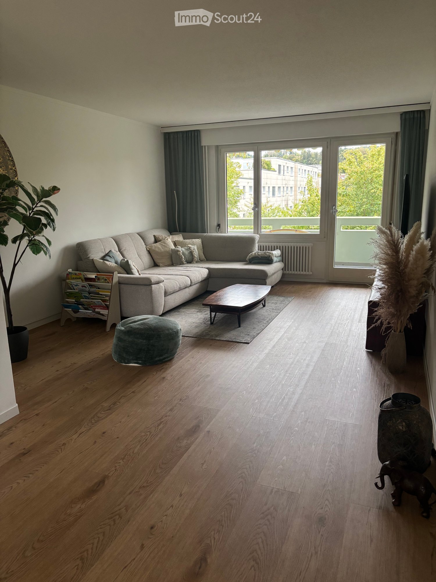 Modern living room with light wooden flooring, large sectional sofa, coffee table, ottoman, decorative vases, and plants.