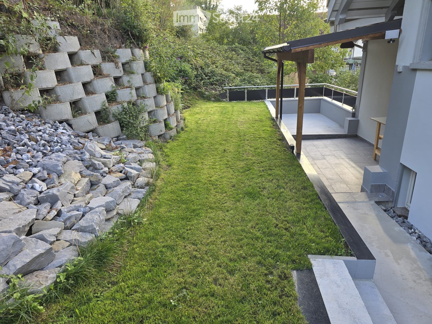 Garden with a grassy lawn, stone retaining wall, concrete steps, and a small patio area with a wooden table.