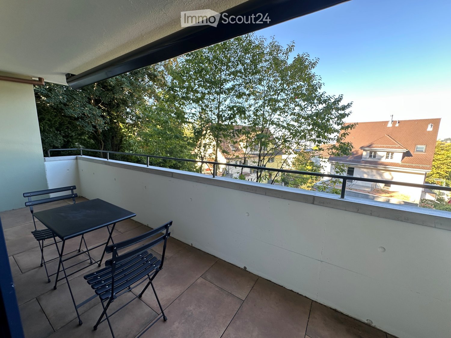 Balcony with tiled floor, white walls, metal table and chairs, metal railing, and view of trees and houses.