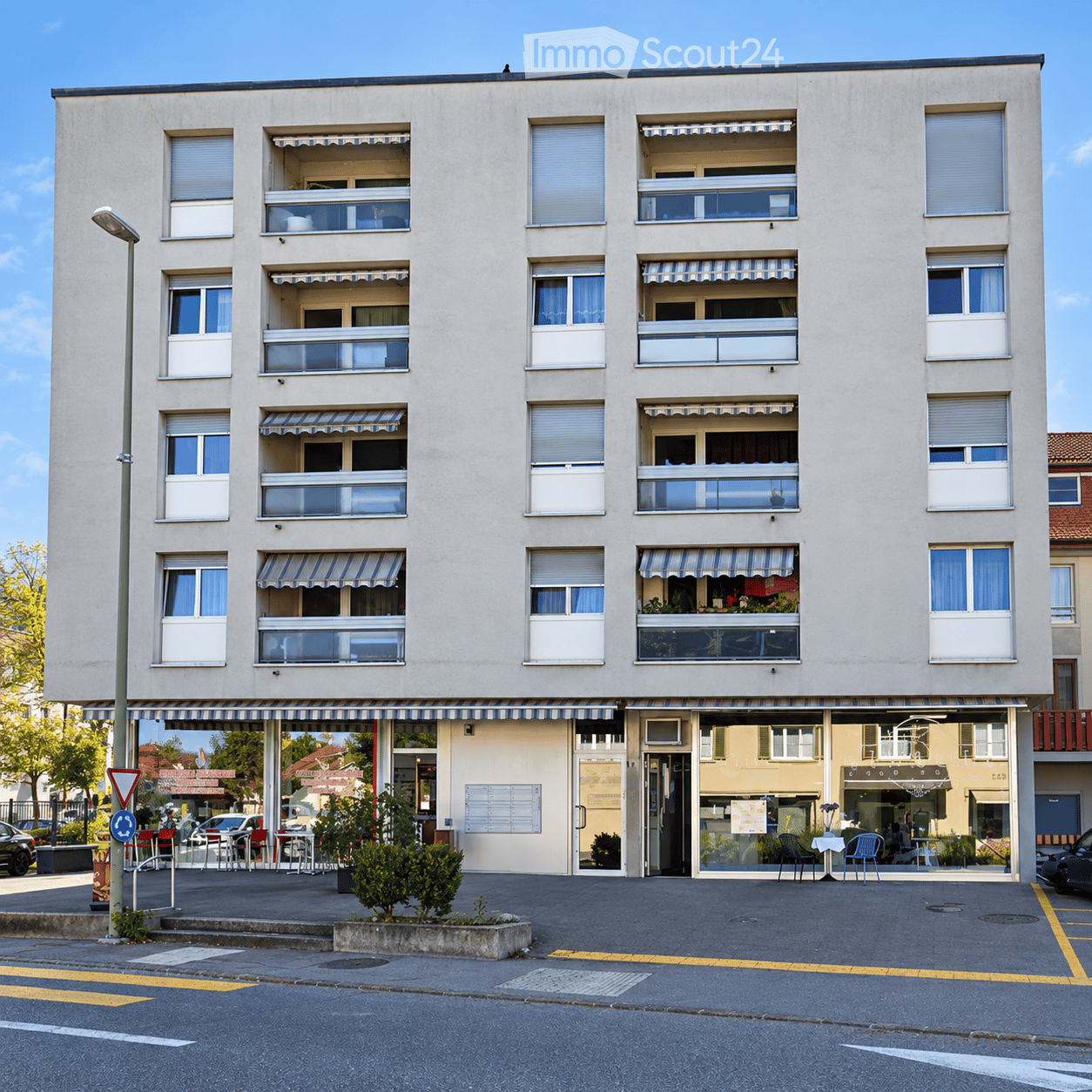 Four-story residential building with multiple balconies, striped awnings, and a storefront at the ground level. Windows have white frames and curtains.