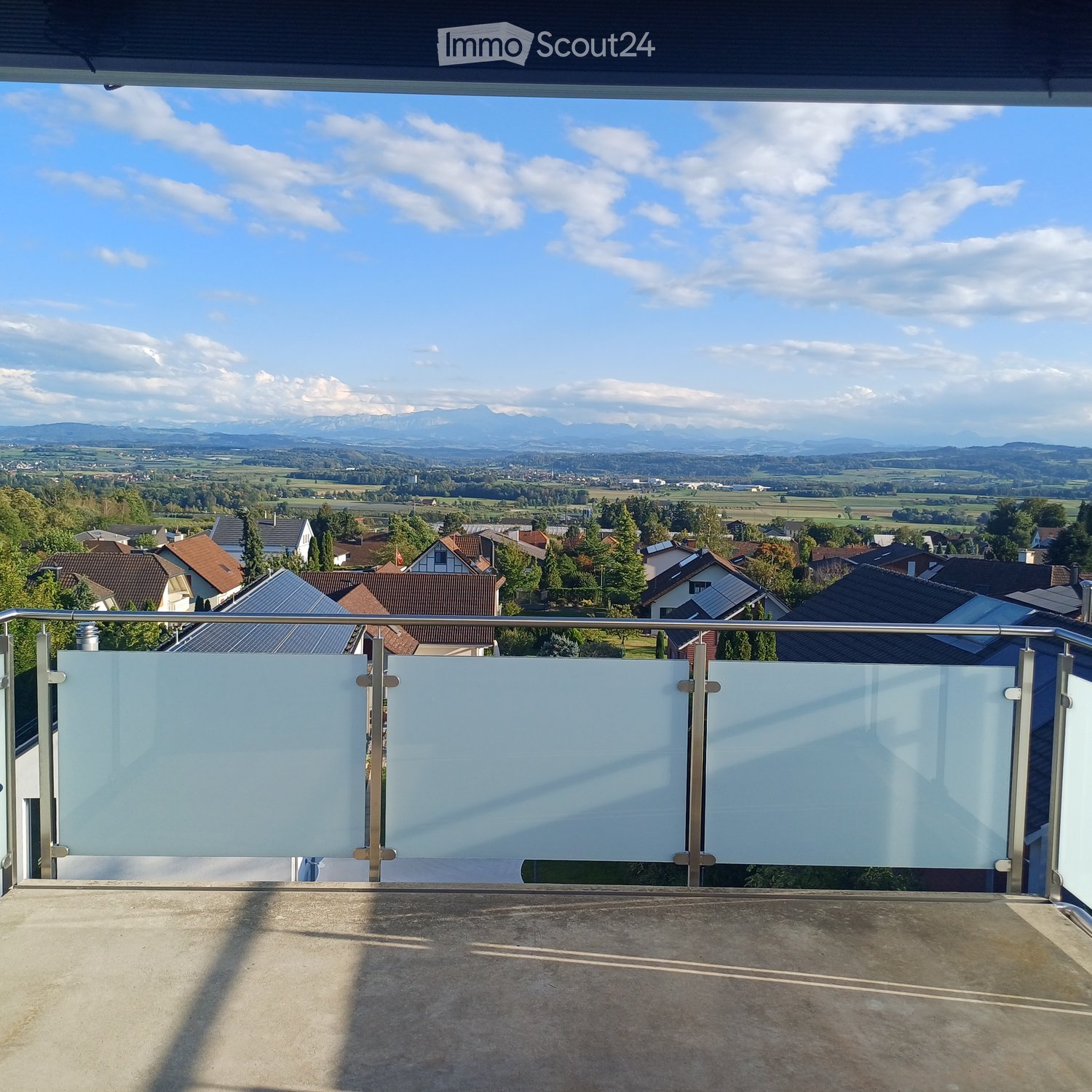 Balcony with glass railing, overlooking a town and distant mountains under a partly cloudy sky.