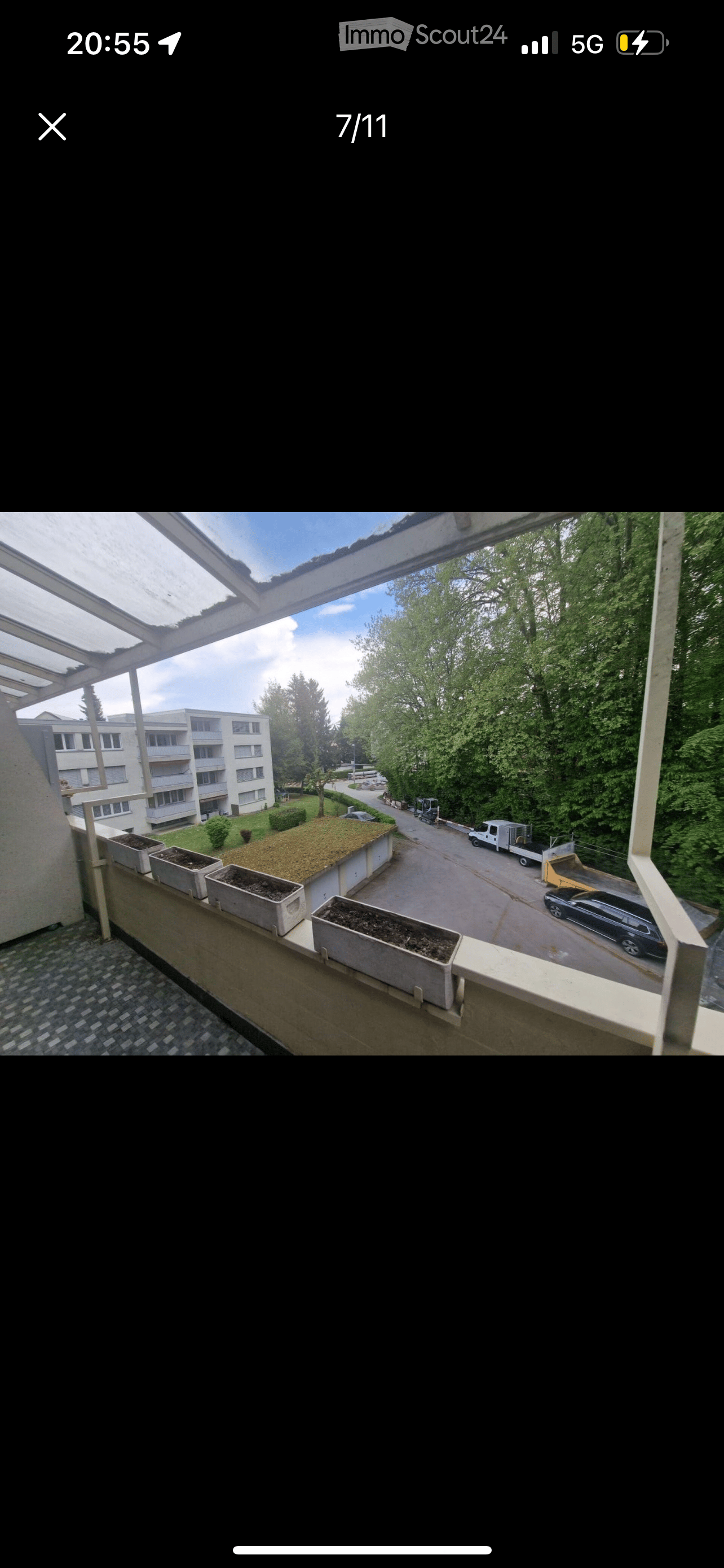 Balcony of an apartment building, with concrete planters, metal railing, asphalt covered ground, green bushes, trees, multi story building, road, parked cars.